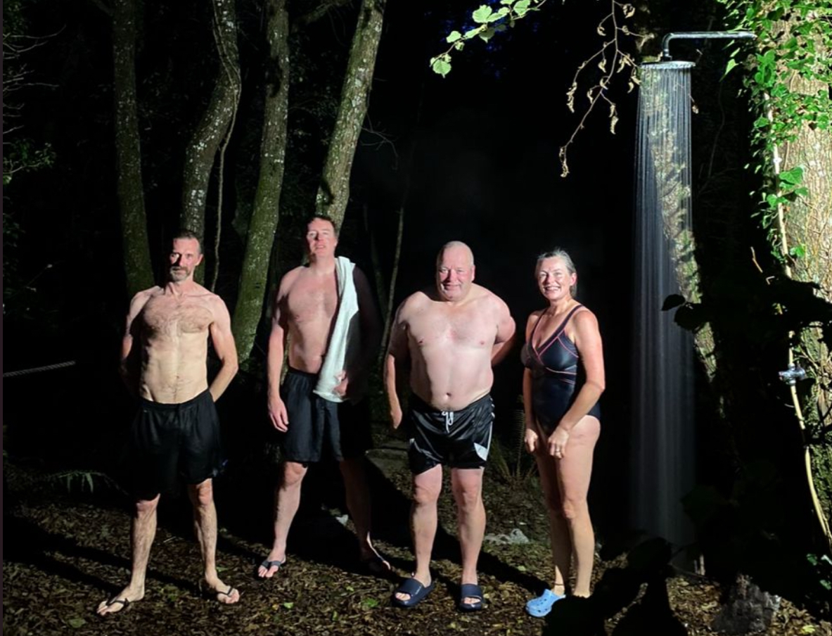 Four adults in swimwear standing outdoors at night near a tree with water flowing from an outdoor shower fixture.