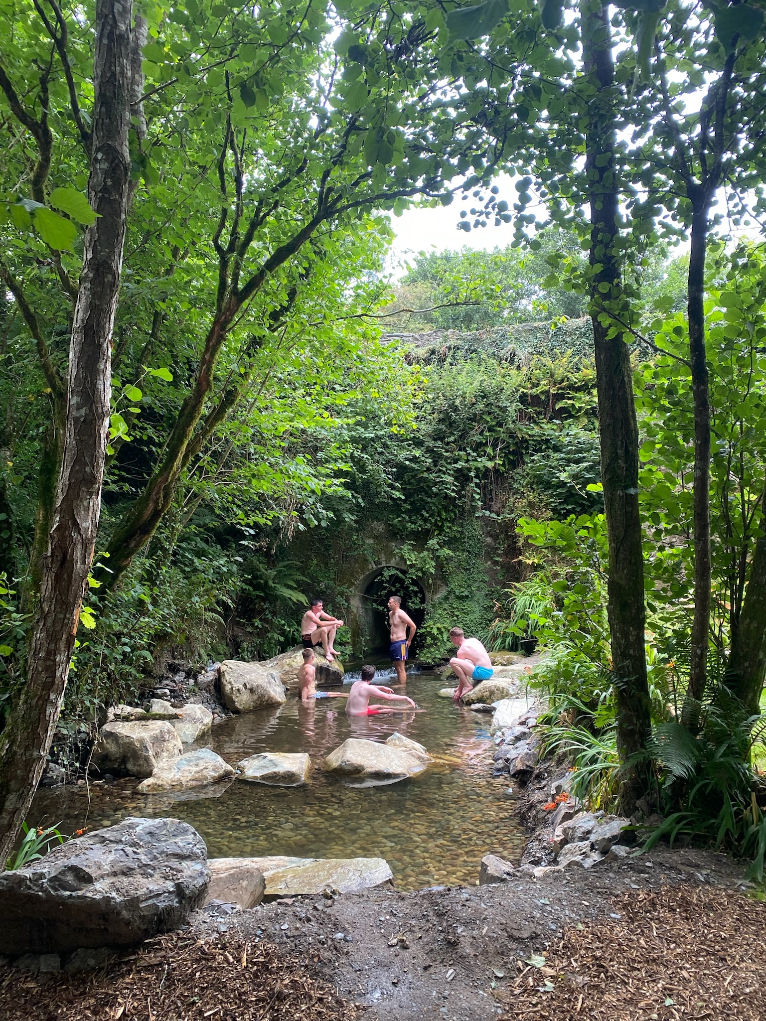 Five people bathing in a shallow stream surrounded by dense green trees and rocks near a tunnel entrance covered in ivy.