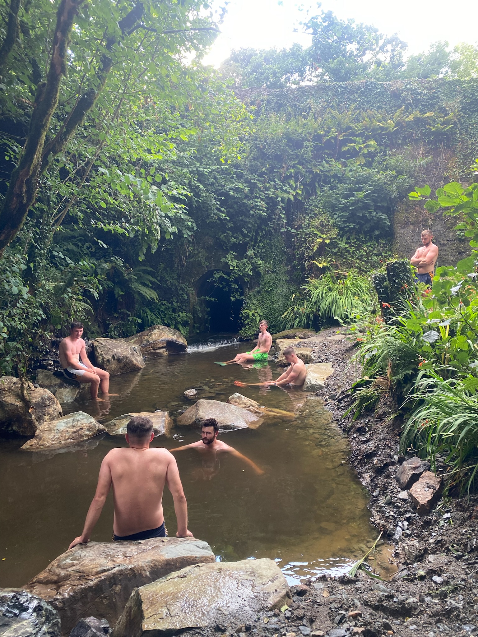 Five shirtless men relaxing and sitting in and around a small natural pool surrounded by rocks and green foliage near a dark cave entrance.