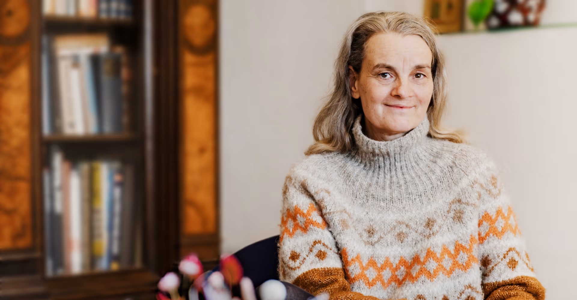 Lächelnde Frau mittleren Alters in einem grau-orange gemusterten Pullover, die drinnen sitzt, mit einem Bücherregal im Hintergrund.
