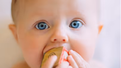 Close-up of a baby with blue eyes holding a wooden block near their mouth.