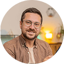 Ein Mann mit Brille und Bart lächelt in die Kamera, trägt ein braunes Hemd, sitzt in einem gemütlichen Raum mit warmer Beleuchtung.