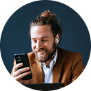 Ein Mann mit Bart im braunen Anzug lächelt, schaut auf sein Smartphone, trägt weiße Ohrhörer.