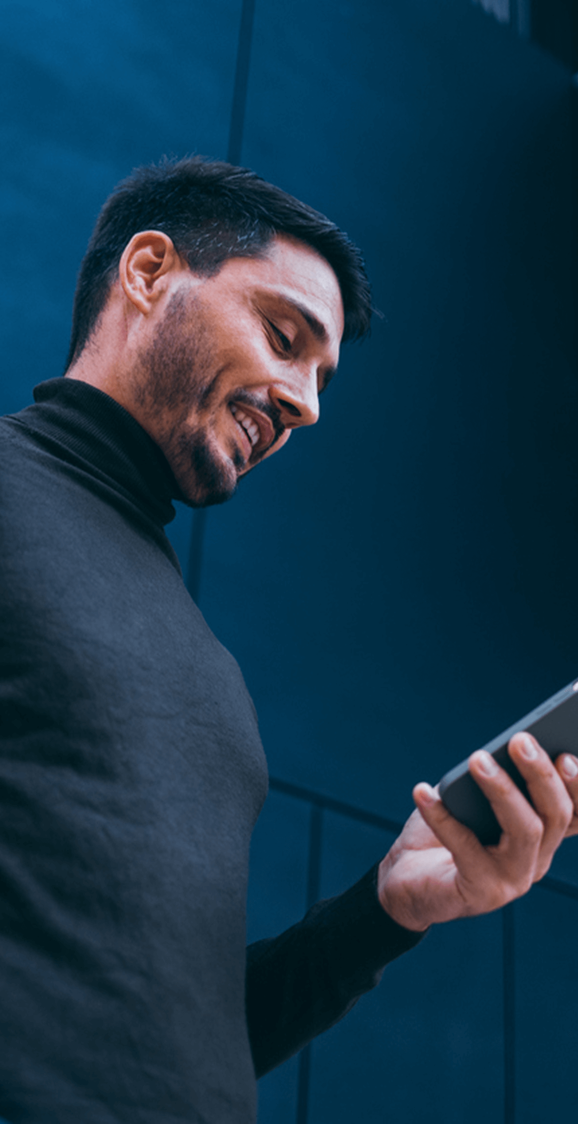 Mann im Rollkragenpullover lächelt auf Smartphone schauend, blauem Hintergrund.