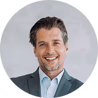 Ein lächelnder Mann in Anzug und Hemd vor neutralem Hintergrund.