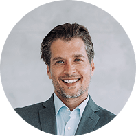 Ein lächelnder Mann in Anzug und Hemd vor neutralem Hintergrund.