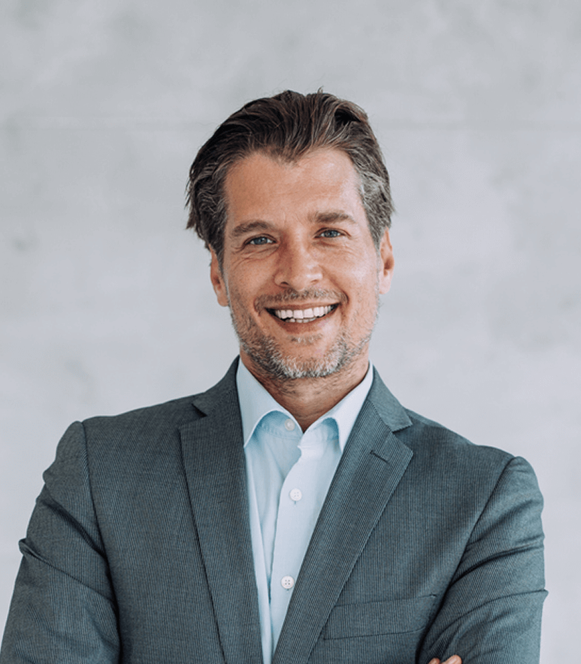 Mann im Anzug lächelt, verschränkte Arme, grauer Hintergrund.