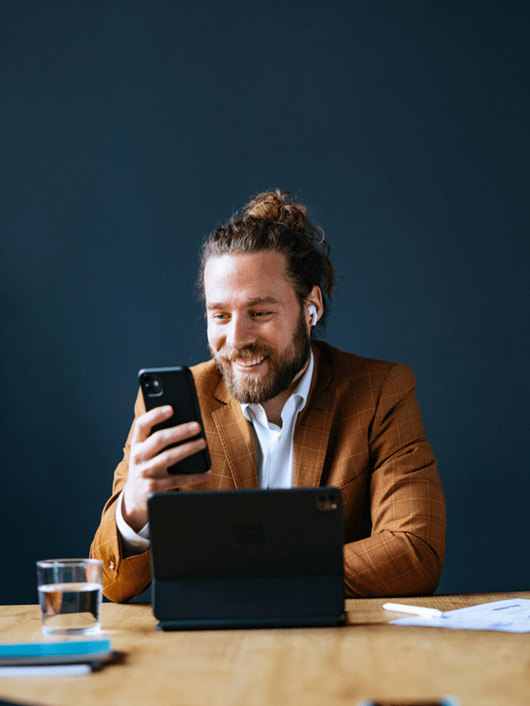 Mann mit Brille und Bart am Schreibtisch, lächelnd, nutzt Smartphone, Tablet vor sich, dunkler Hintergrund.