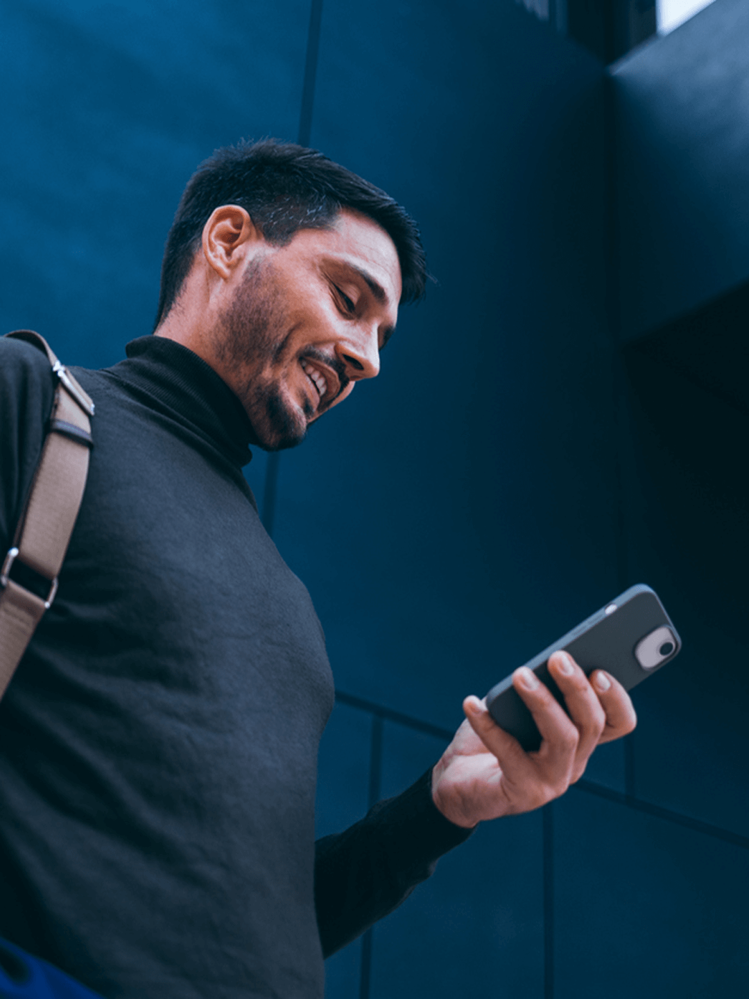 Mann in schwarzem Rollkragenpullover lächelt auf Handy in moderner urbaner Umgebung.