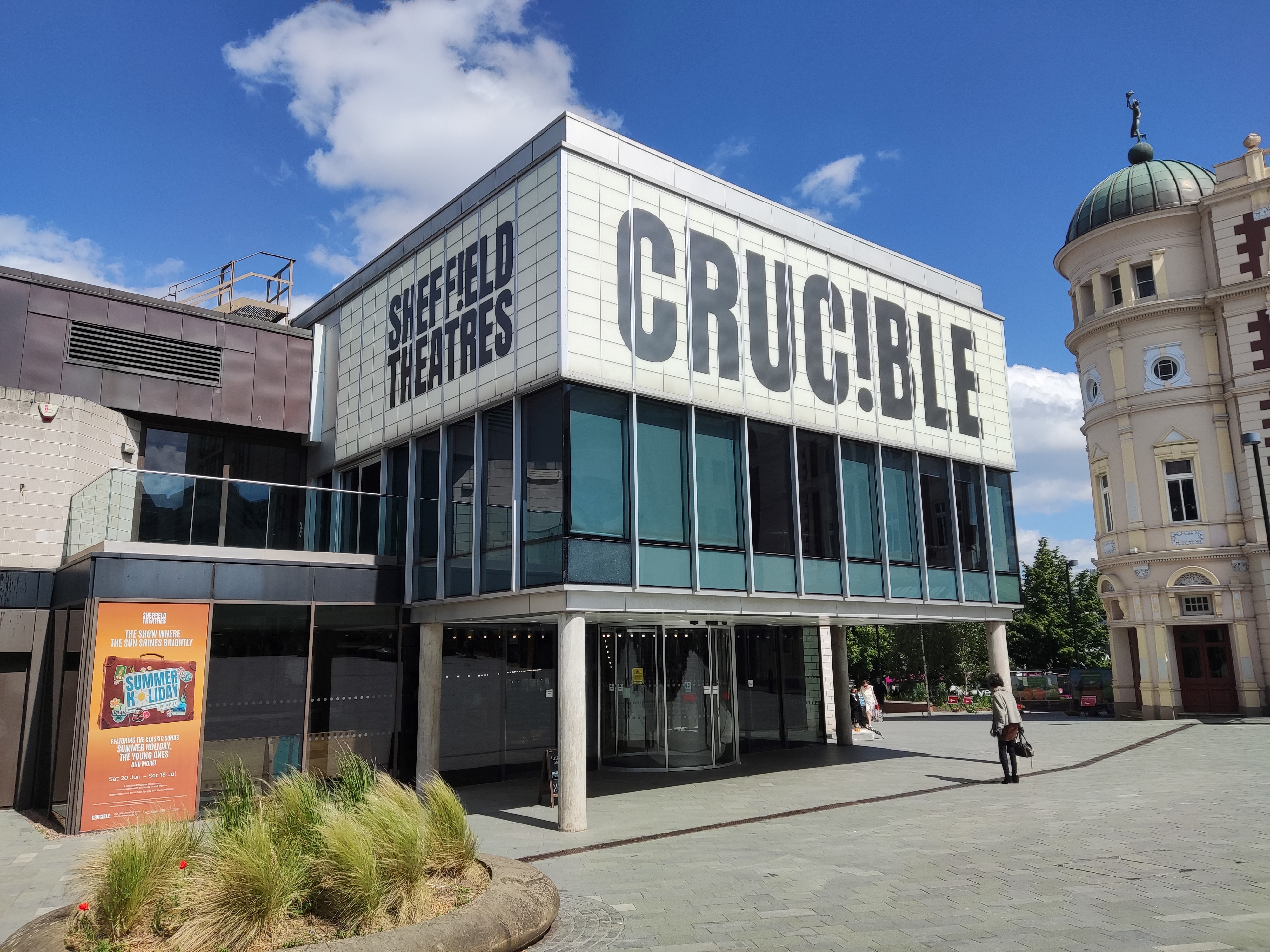 The Crucible Theatre on a sunny day. The building is three storeys tall with a modern, boxy shape and large signs covering the third floor reading "Sheffield Theatres" on the side, and "Crucible" on the front.