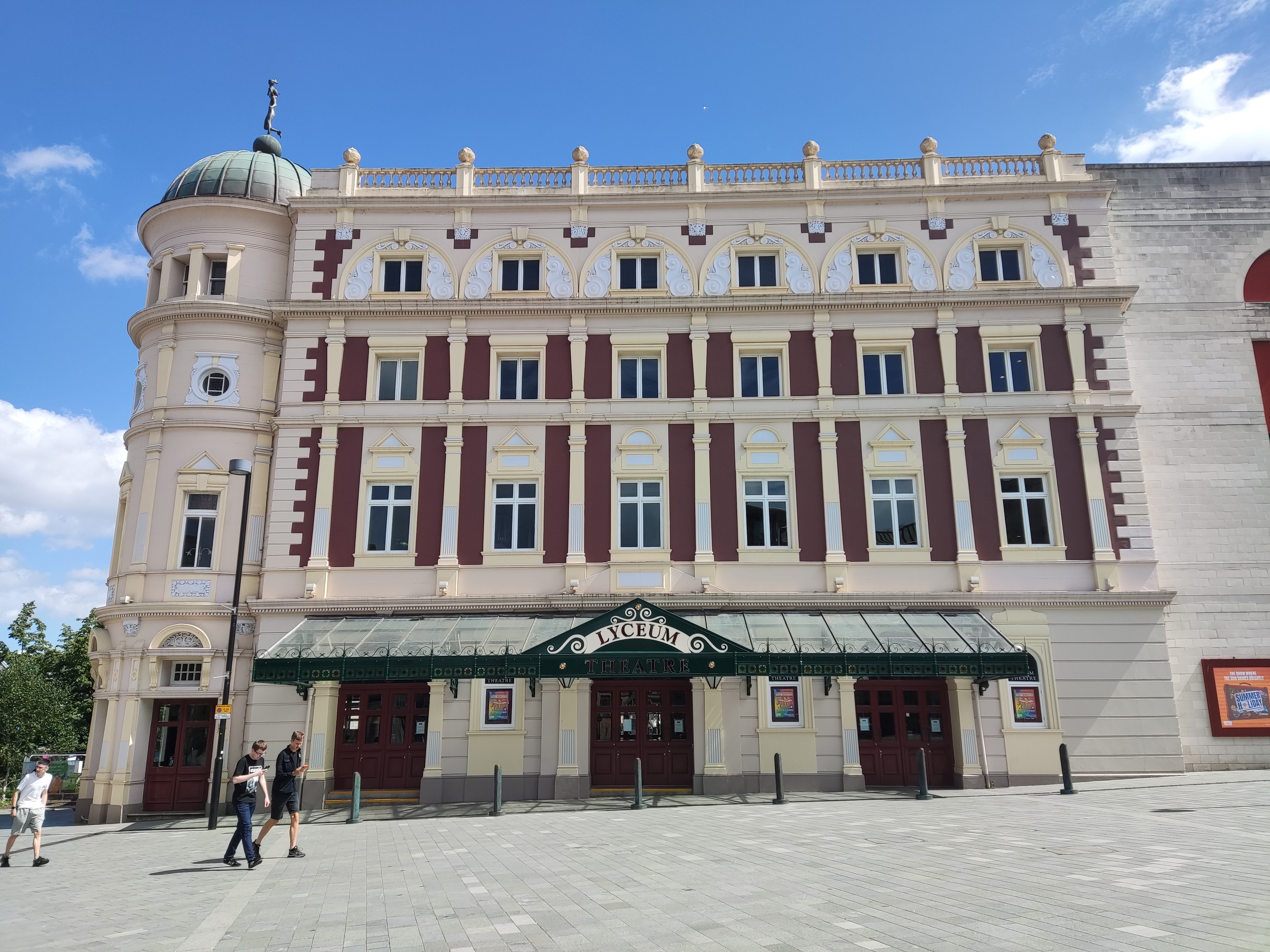 The main facade of the Lyceum Theatre on a sunny day. The theatre is four storeys tall with a domed turret on the left and a mix of decorations including an ironwork awning, decorative columns, roof balustrade and a statue atop the dome.