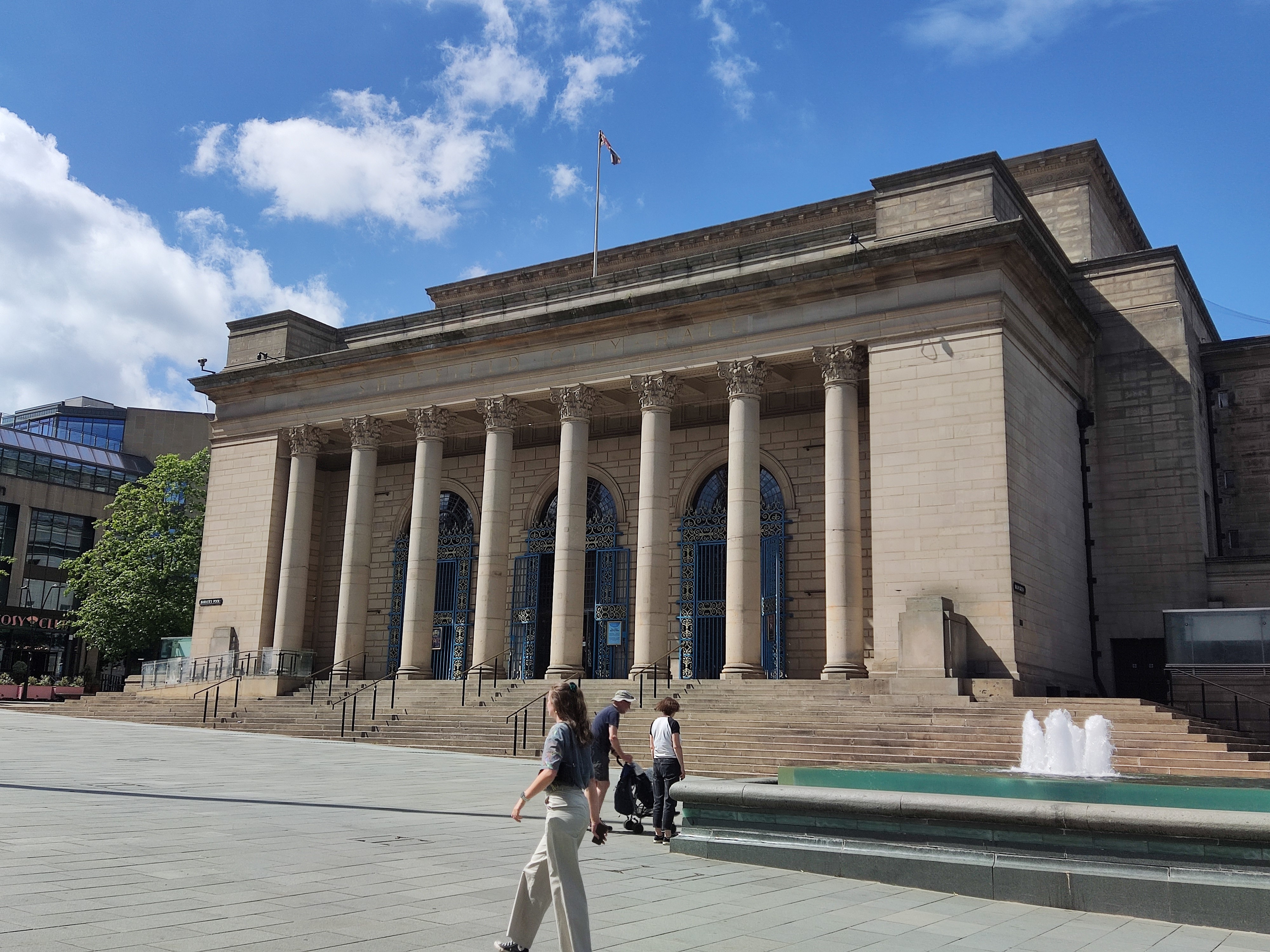 Sheffield City Hall on a sunny day. The grand, neo-classical stone building features eight large corinthian pillars across its centreal, flat-roofed portico topped by a flagpole and further tiers of flat roofs behind