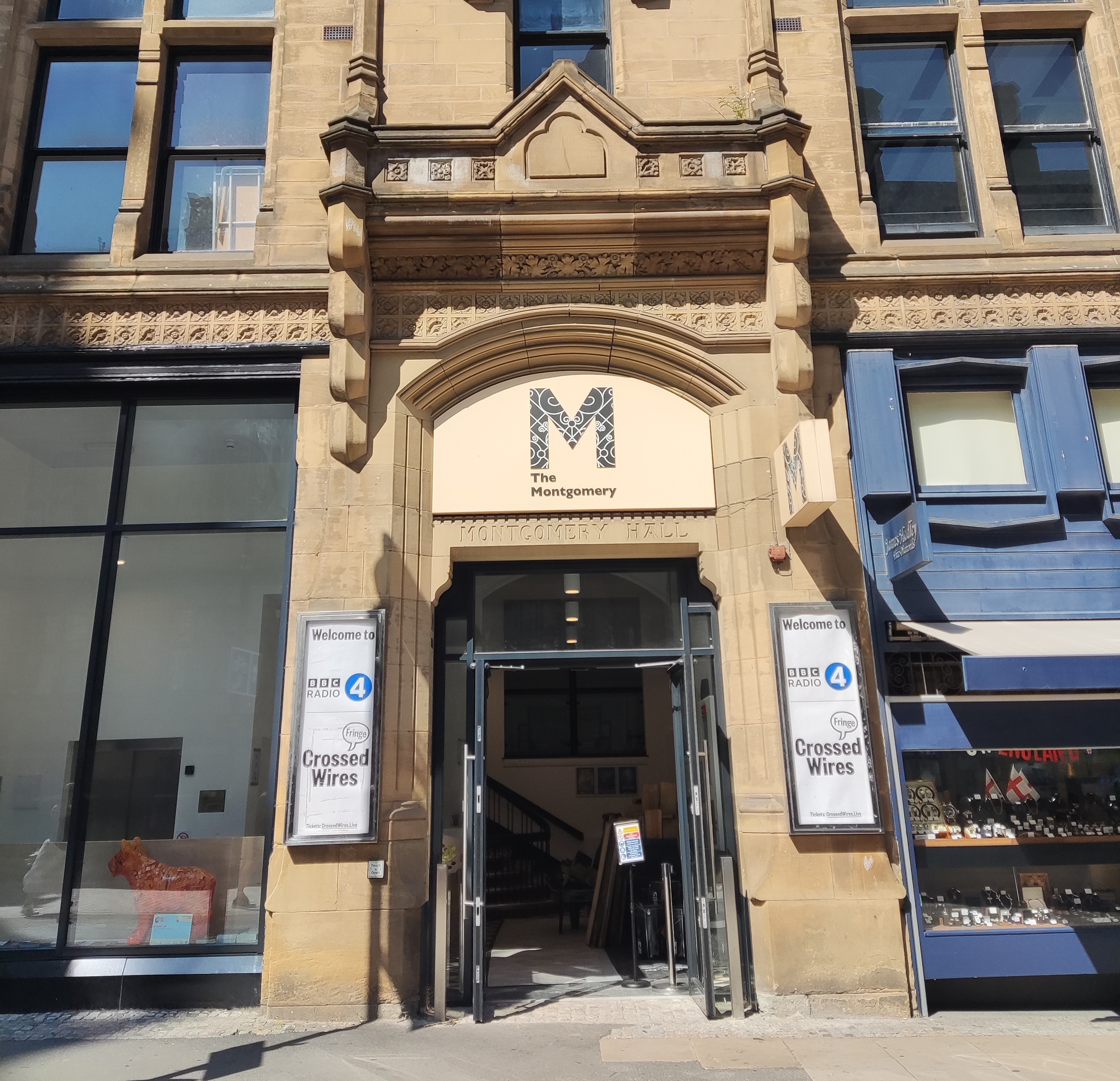 Sheffield's Montgomery Theatre on a sunny day. The stone entrance is topped by a decorative, engraved entryway and features glass doors, a sign above with the theatre's name and signs either side advertising their current events.