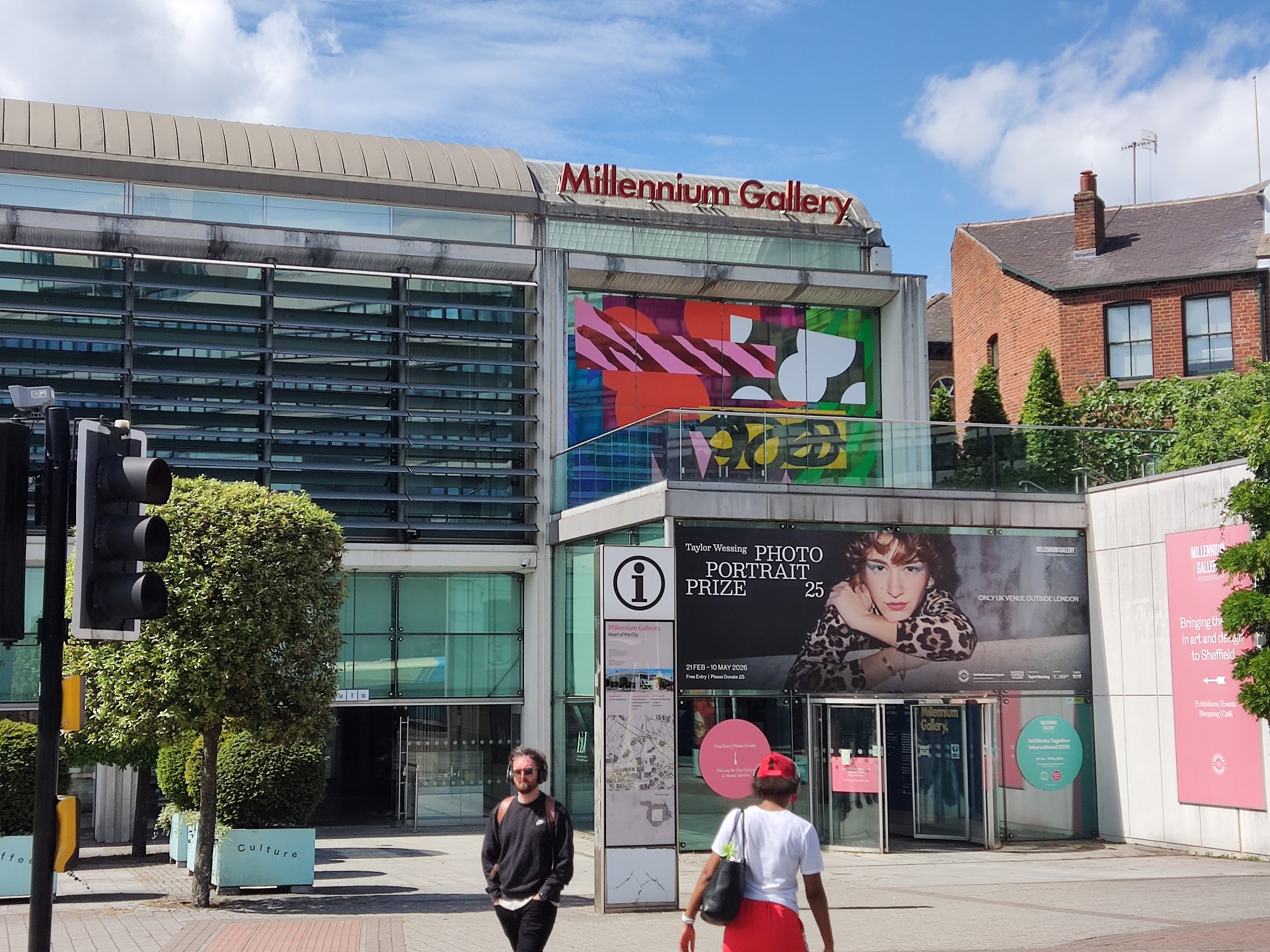 The Millennium Gallery on a sunny day. The gallery's light, glass-covered facade includes colourful, patterned window decortions, a sign advertising a portrait exhibition and the another with the gallery's name.