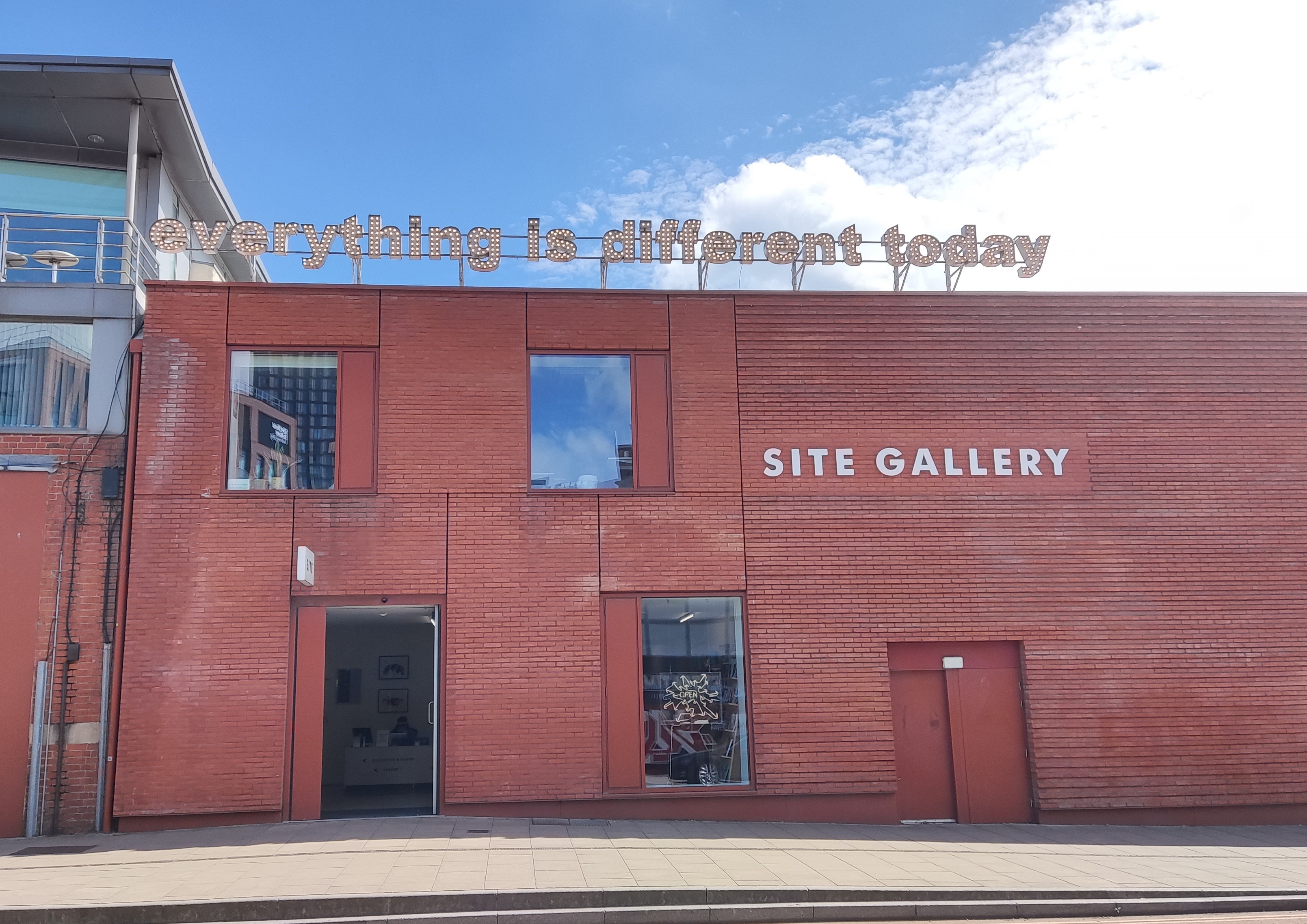 The Site Gallery on a sunny day. The two-storey building's orange brickwork is textured in parts and supports a sign with the gallery's name. Additionally, above the flat roof, a light-up sign reads "everything is different today".