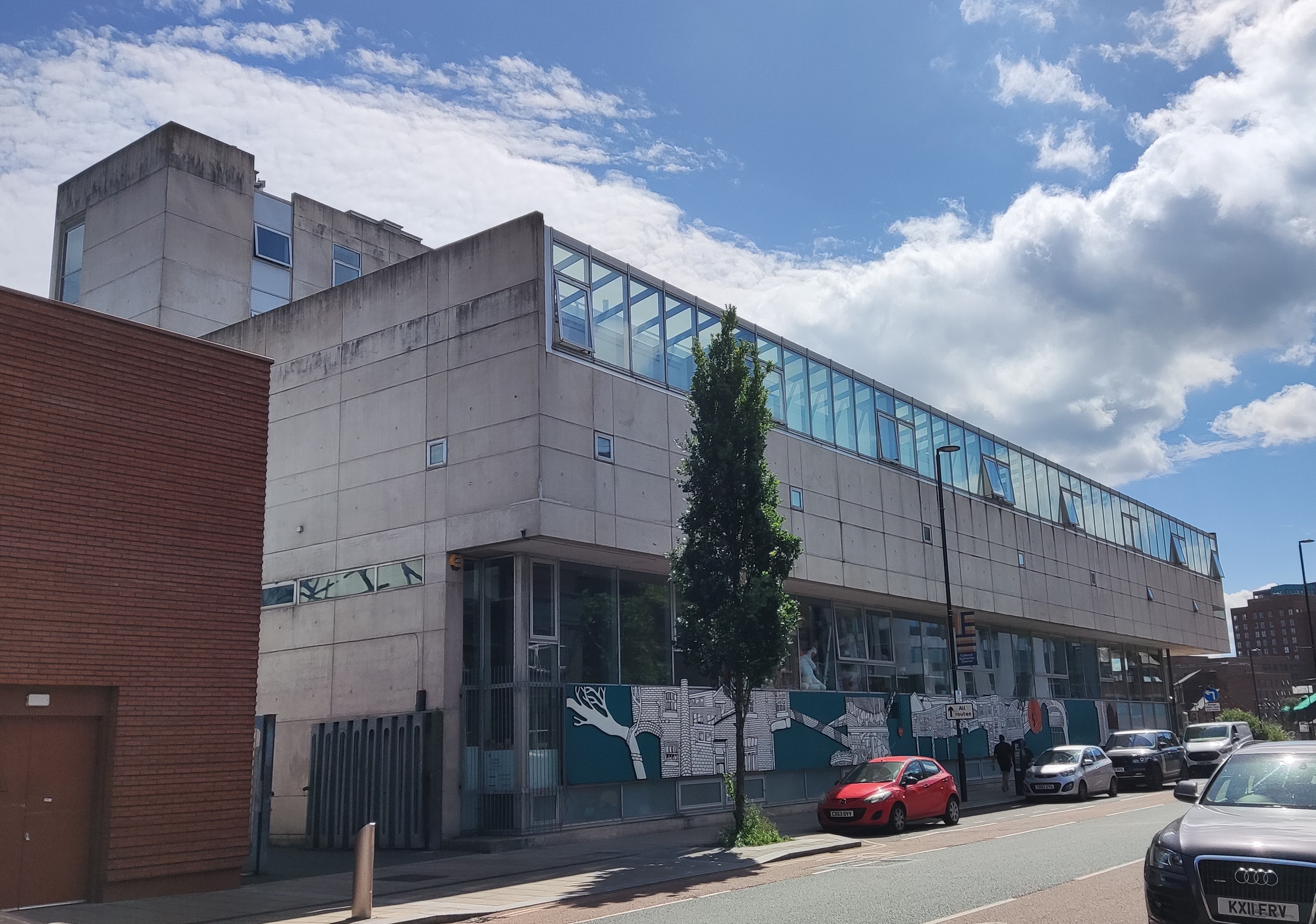 The Yorkshire Art Space on a sunny day. The building's brutalist and blocky concrete exterior is softened by a row of windows and skylights across the front that serve the artists' studios. Below, on the ground floor, a long mural of sheffield buildings provides visual interest to passersby.