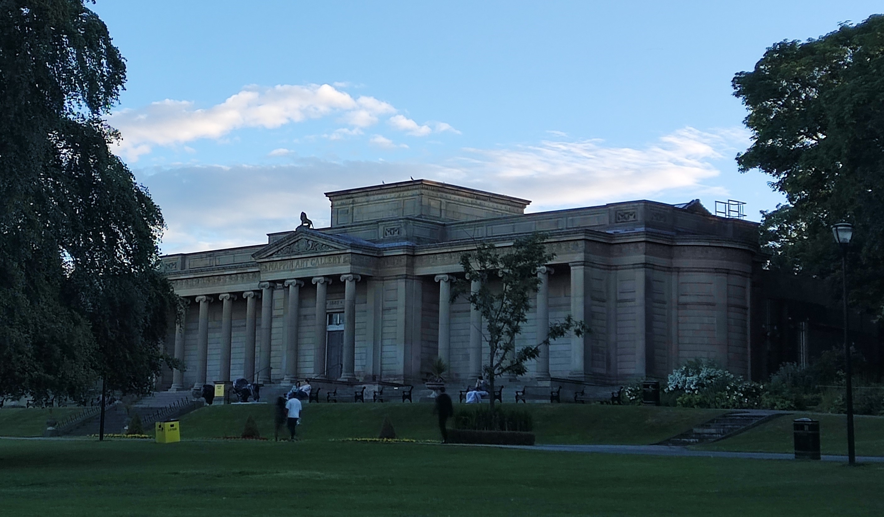 Weston Park Museum at dusk. The neo-classical stone building supports a row of ionic columns aross the wings and front portico with a lion statue standing in the centre of the roof.
