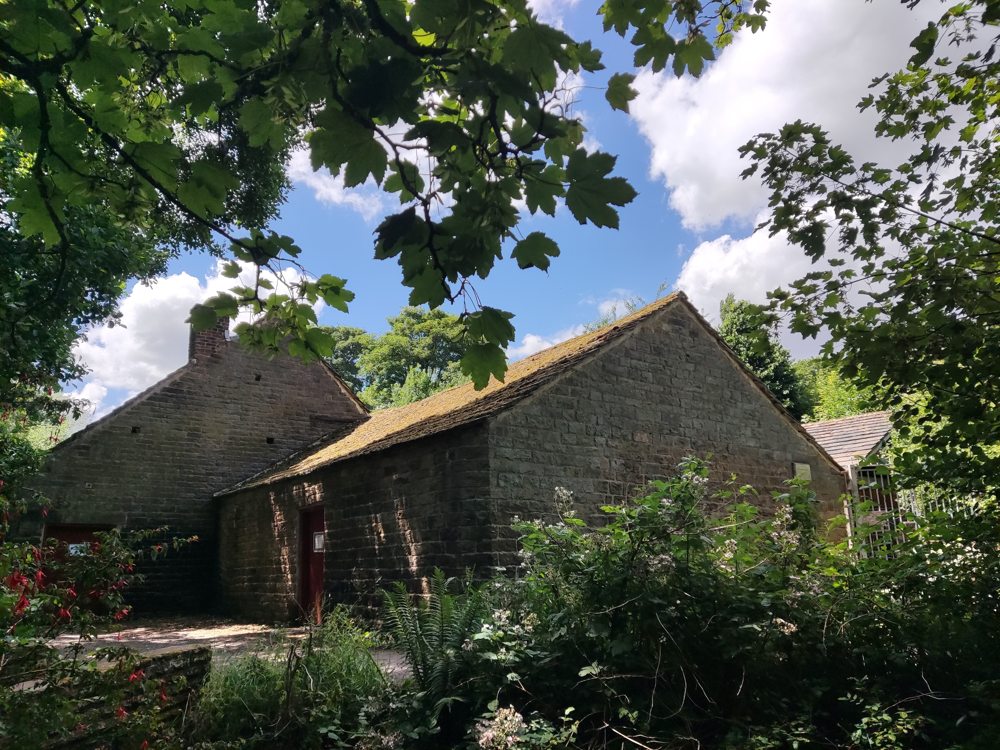 Shepherd Wheel on a sunny day surrounded by the green foliage of bushes and trees. The low, stone buildings have mossy slate roofs, no visible windows, and are accessed by a small footbridge across the Porter River.