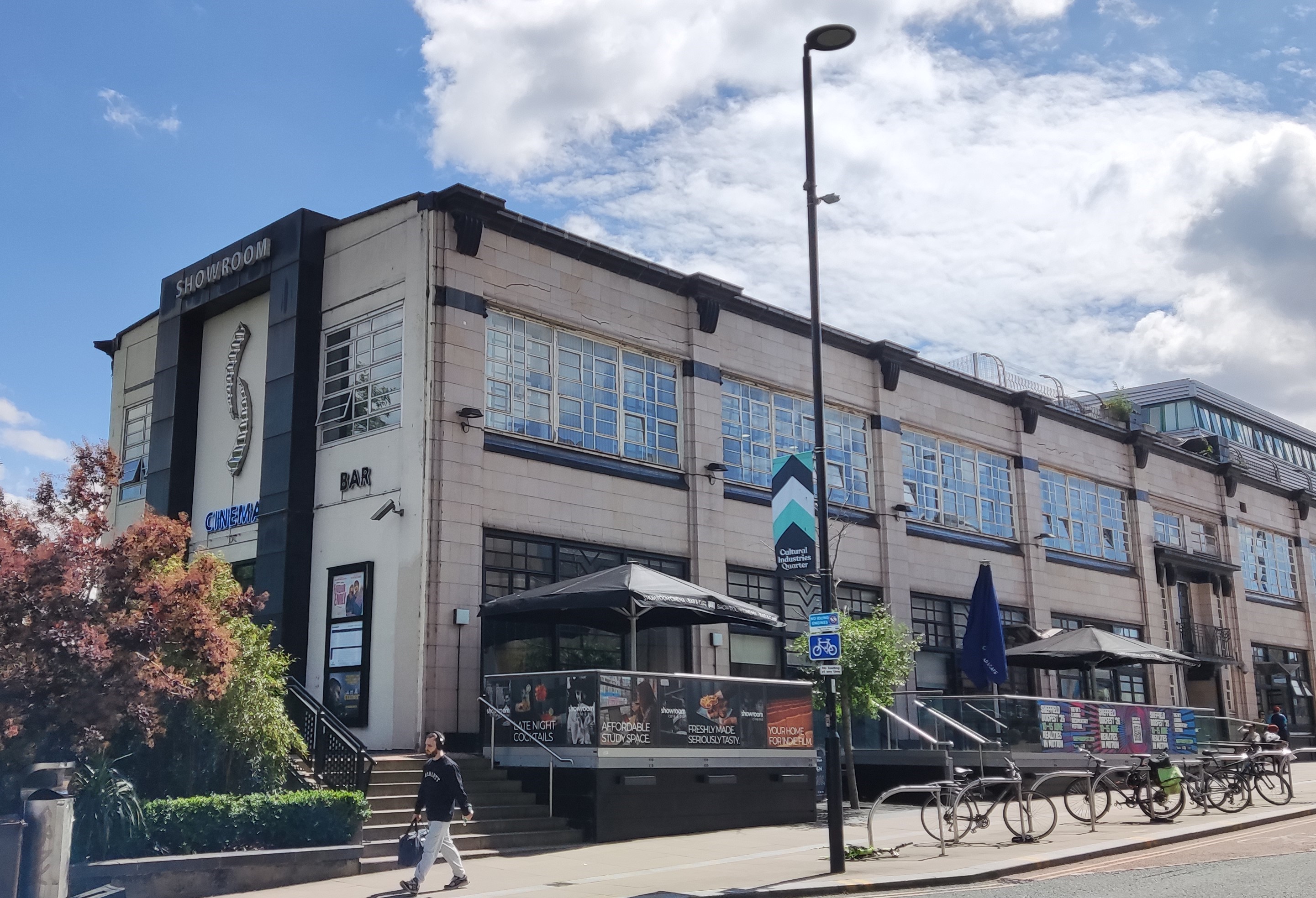 The Showroom cinema on a sunny day. The art-deco building stands two storeys tall and is faced with white tiles and black accents around the roof, windows and main entrance. The Showroom's cafe terrace can also be seen overlooking the road with umbrellas raised above the tables.