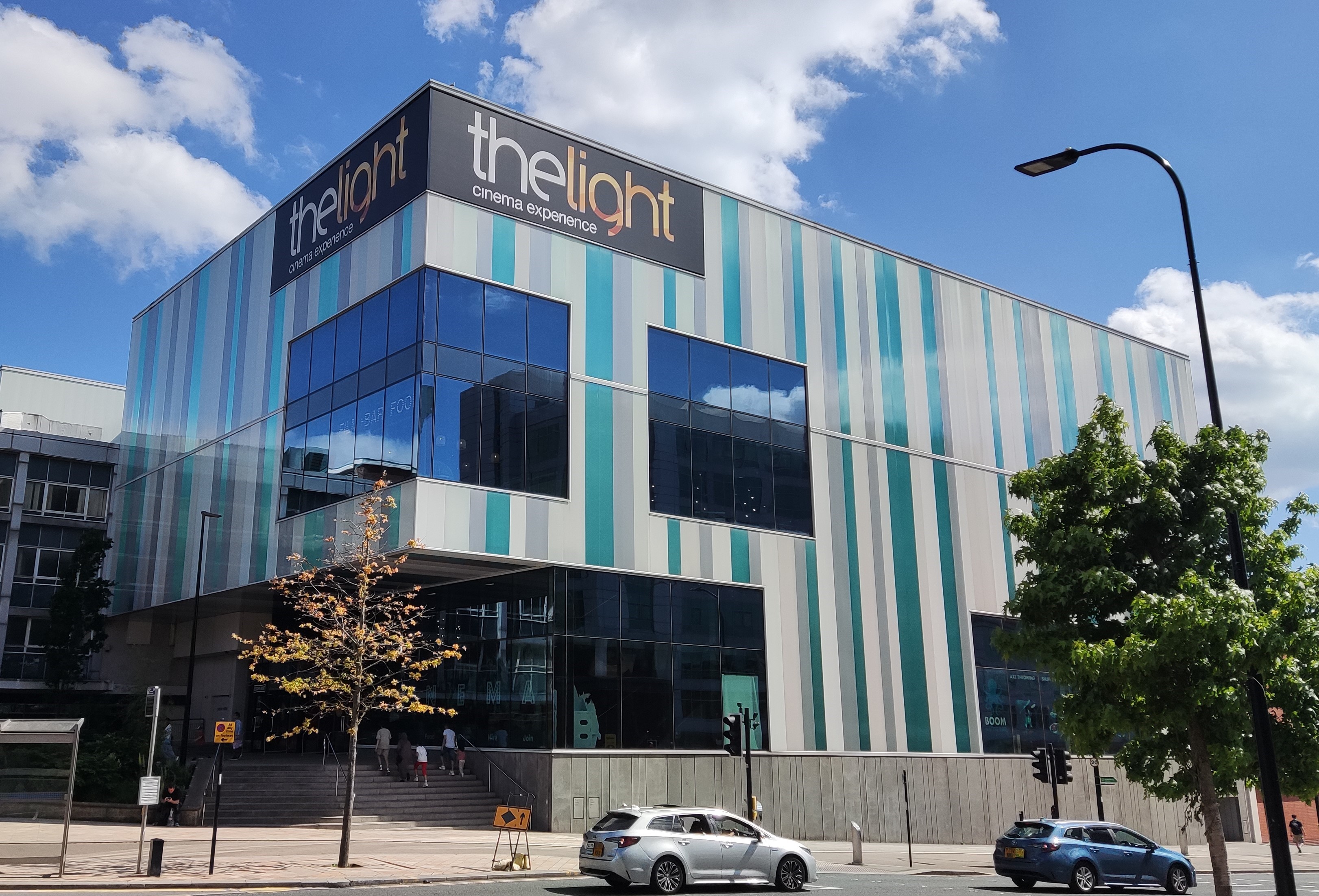 The Light Cinema on a sunny day. The four storey, rectangular, steel and glass building has two big windows on its left hand side beneath a sign with the cinema's name. Below, steps rise up to the main entrance.