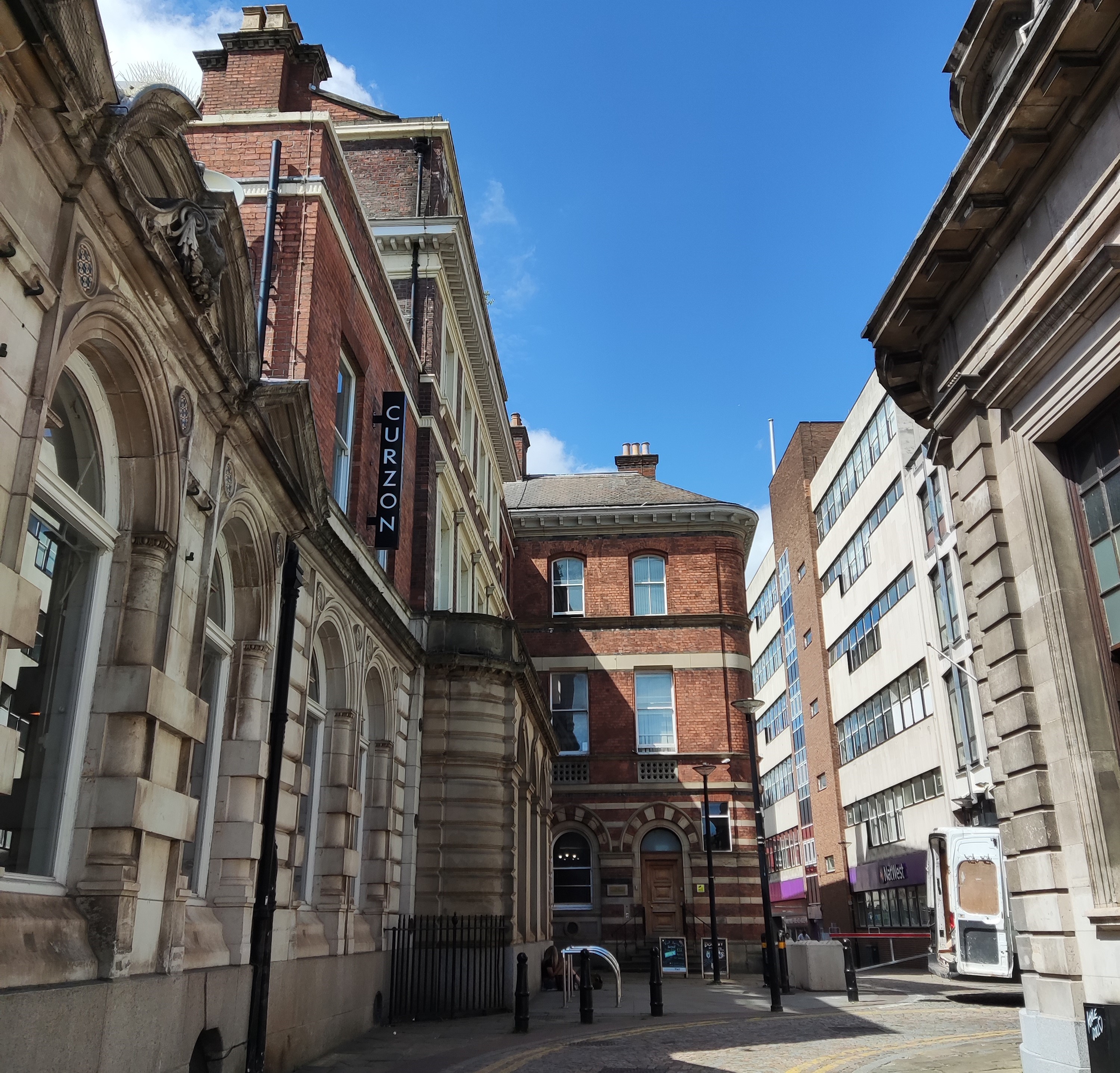 The Curzon Cinema on a sunny day. The four-storey noeclassical/ Georgian building rises to the left of the thin street in brick and stone.