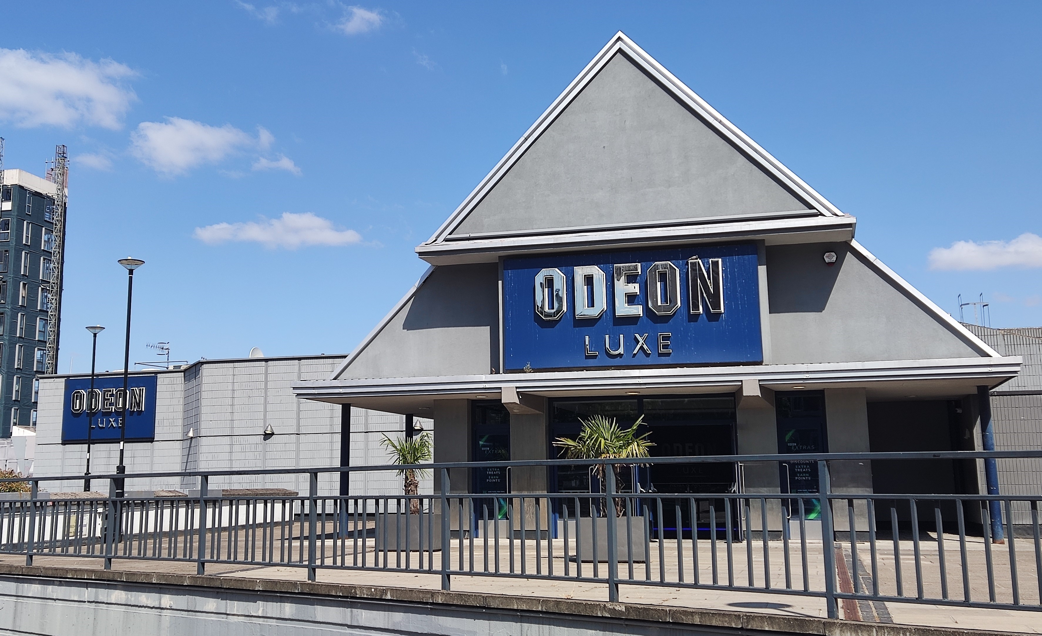 The Odeon cinema on a sunny day. The large, grey building is fronted with a triangular portico supporting a large blue sign that reads "Odeon Luxe" in Chrome letters.