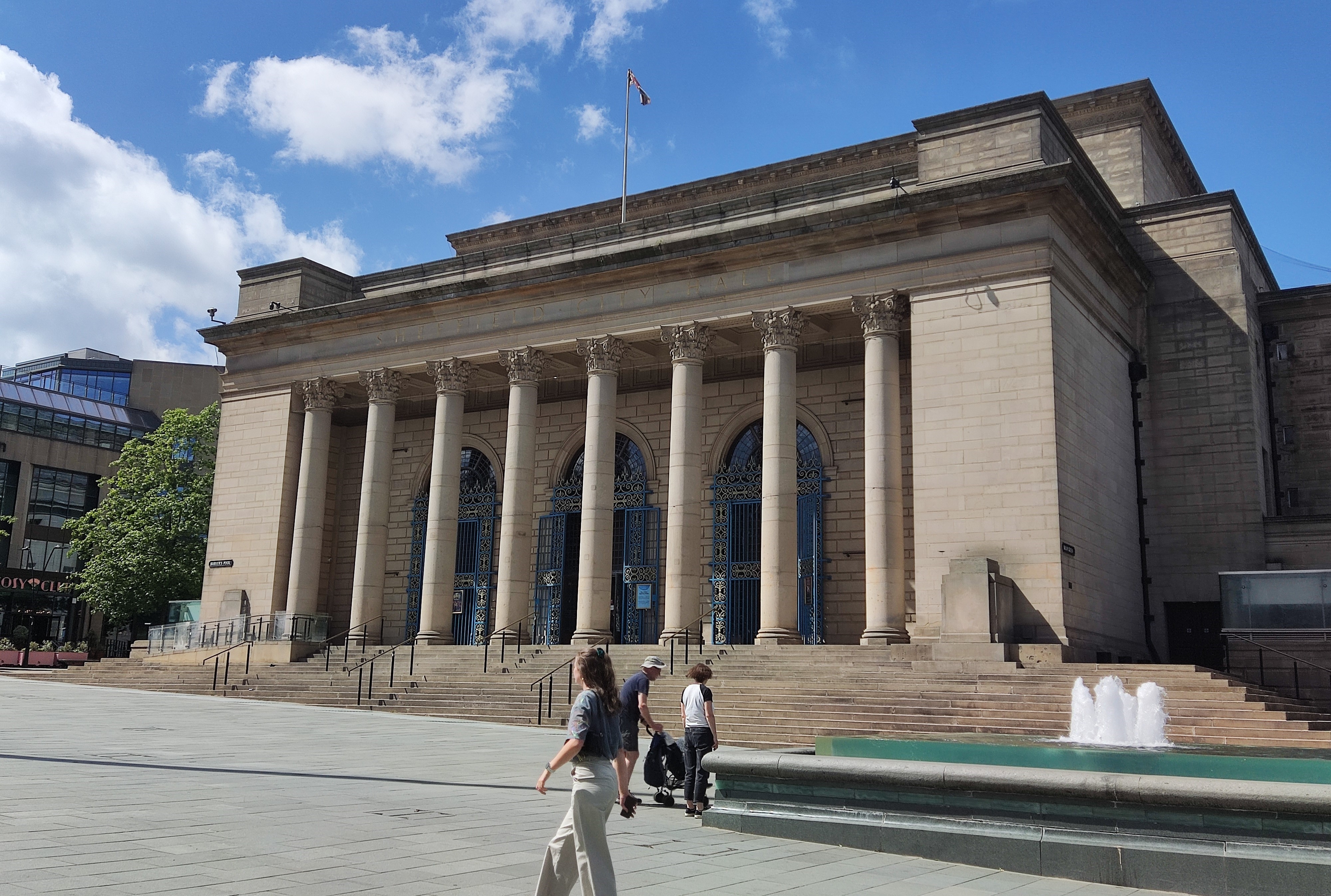Sheffield City Hall on a sunny day. The grand, neo-classical stone building features eight large corinthian pillars across its centreal, flat-roofed portico topped by a flagpole and further tiers of flat roofs behind.