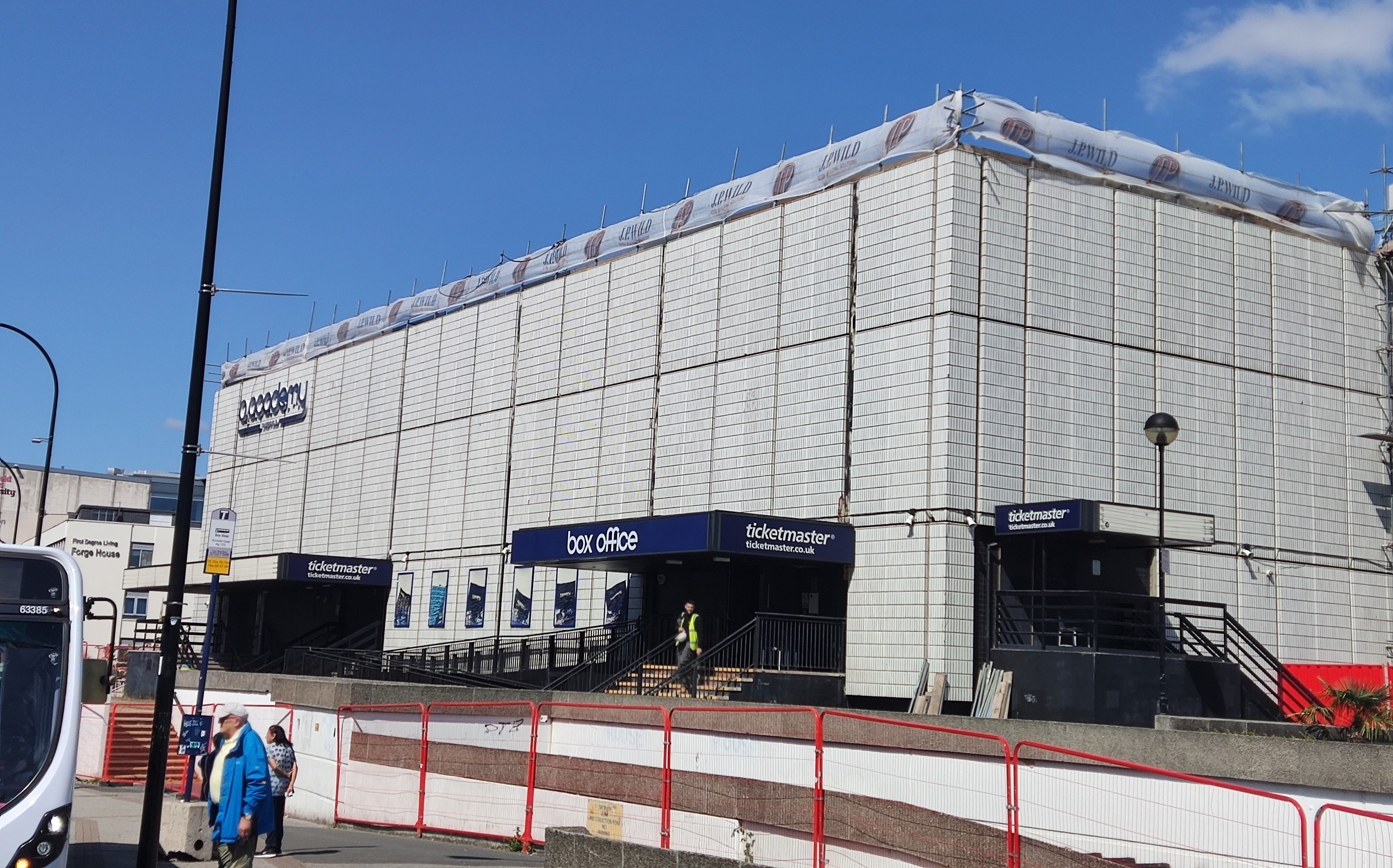 Sheffield's O2 Academy on a sunny day. The white-tiled, blocky, 3 storey building is pictured at an angle, showing the "O2 Academy" and "box office" signs in blue near the roof and the entrance respectively.