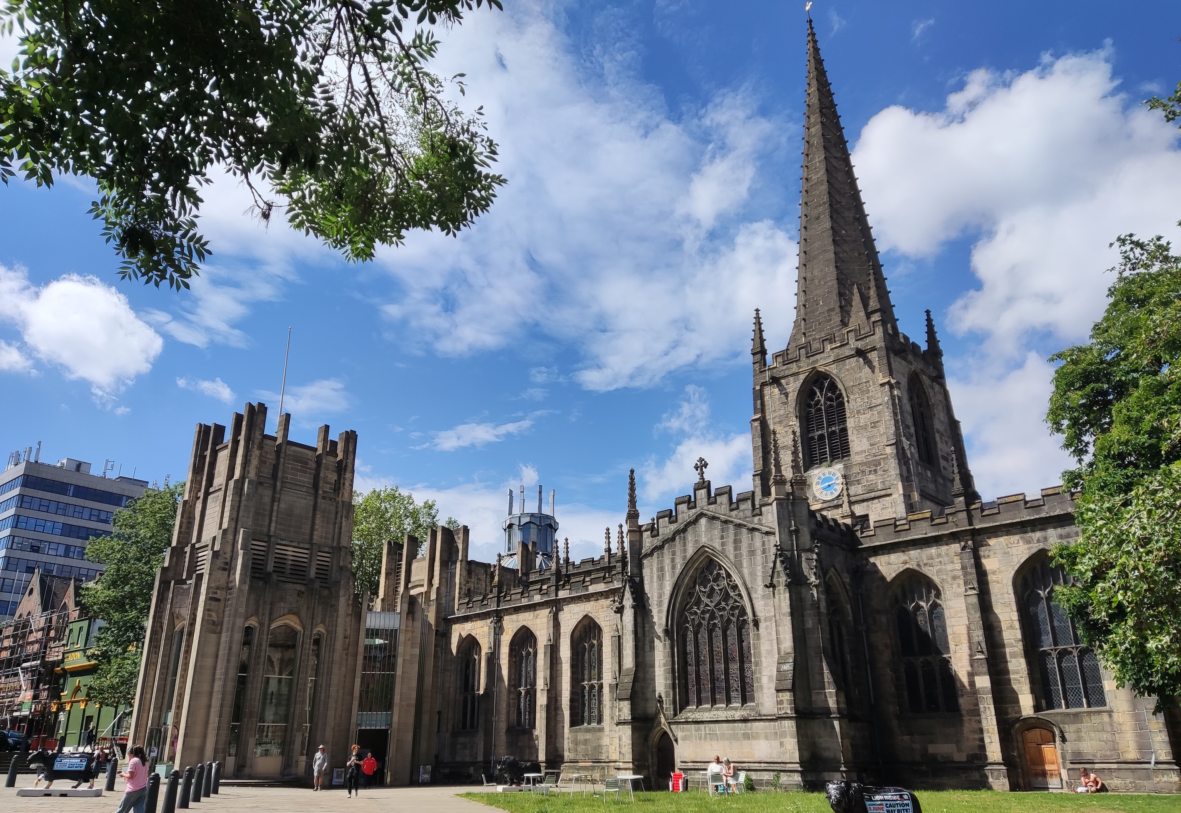 Sheffield Cathedral on a sunny day. The gothic stone cathedral features grand, vaulted windows, a central masonry spire and additional, shorter towers added later.