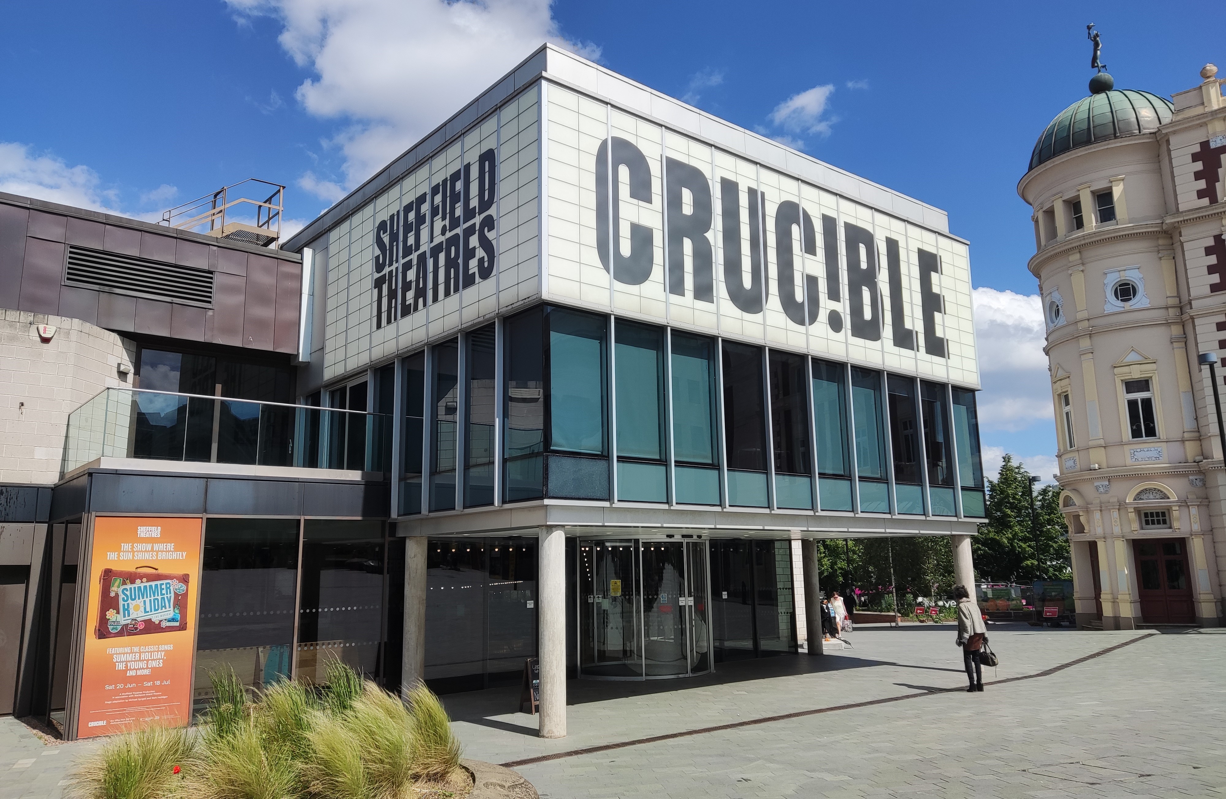 The Crucible Theatre on a sunny day. The building is three storeys tall with a modern, boxy shape and large signs covering the third floor reading "Sheffield Theatres" on the side, and "Crucible" on the front.