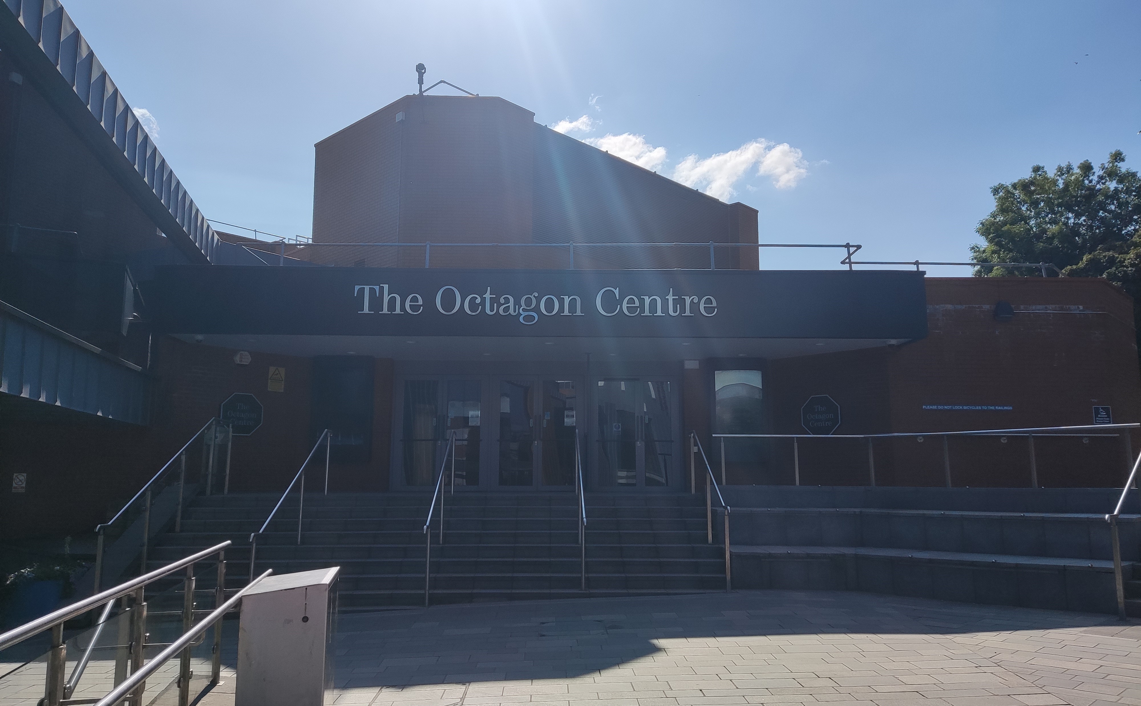 The Octagon Centre on a sunny day. A flight of steps leads up to the venue's glass doors which stand beneath a blue with the venue's name. The venue is one story tall and built of red brick.