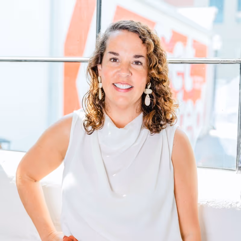 Smiling woman with curly hair wearing white sleeveless top and statement earrings, sitting indoors by a window.