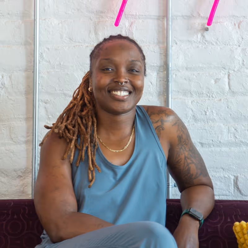 Smiling person with dreadlocks, tattoos, and nose rings sitting and wearing a blue tank top.