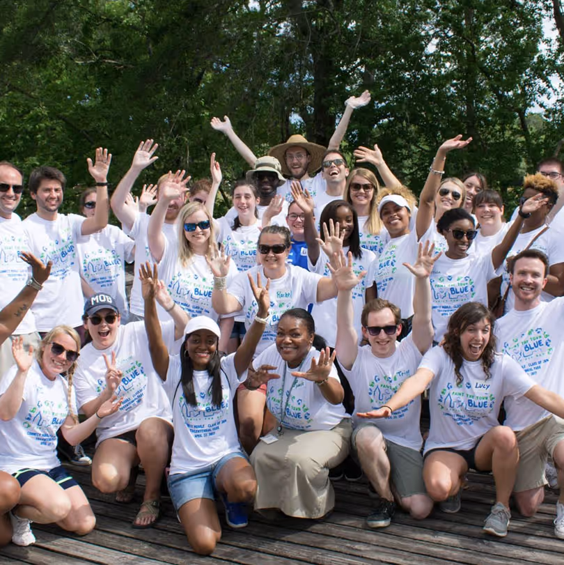 Group of diverse people wearing matching white event t-shirts smiling and waving outdoors on a wooden deck with trees in the background.