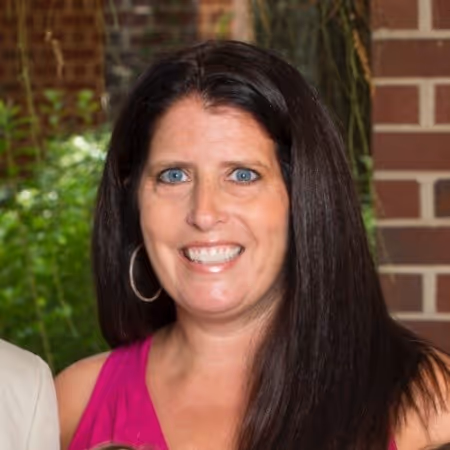 Smiling woman with long dark hair wearing a magenta sleeveless top and hoop earrings, standing in front of a brick wall with greenery in the background.