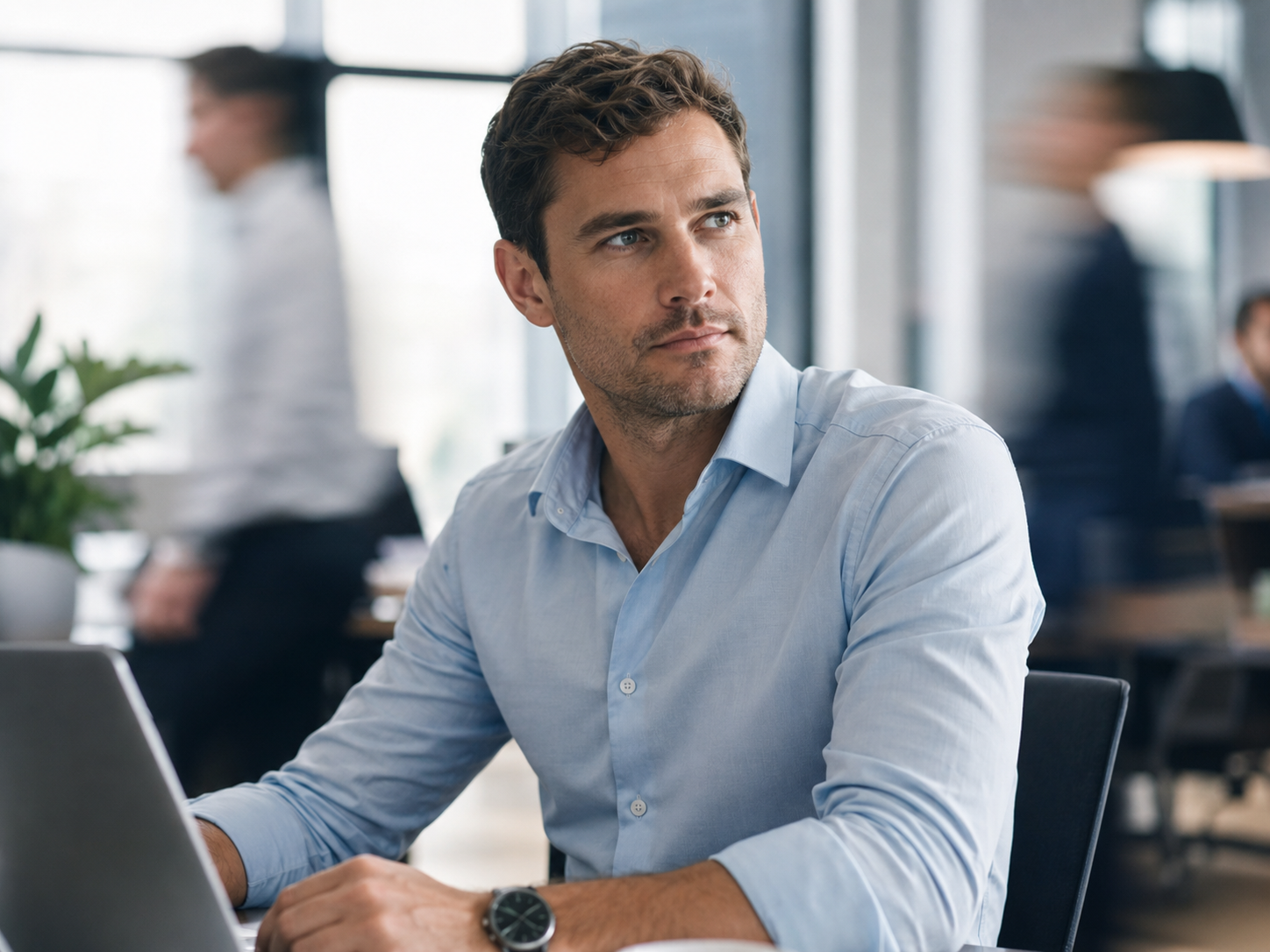 Man in light blue shirt sitting at desk with laptop in a busy office, looking thoughtfully to the side.