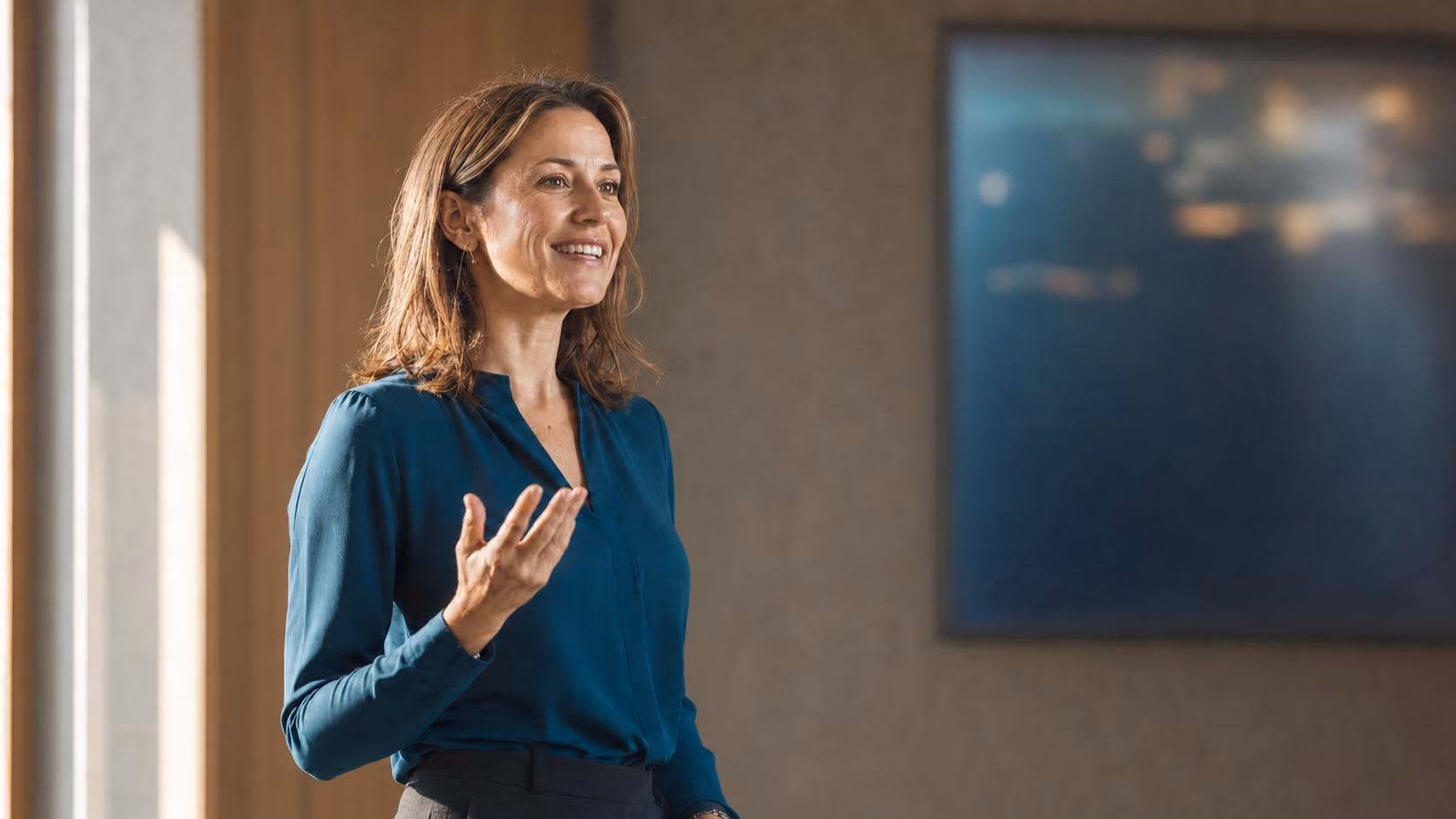 Smiling woman in teal blouse speaking and gesturing with hand indoors against a blurred background.