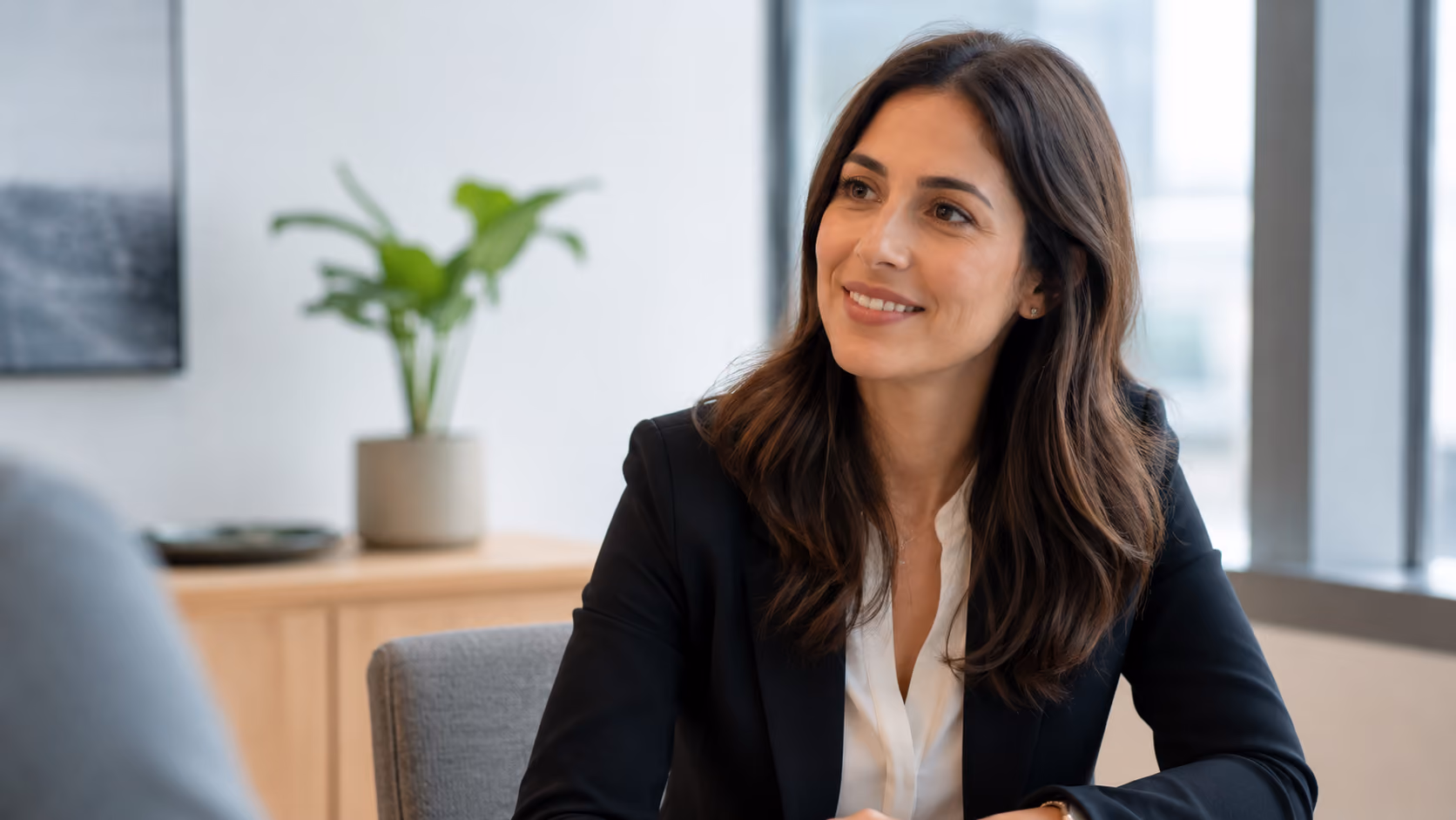 Smiling woman with brown hair in black blazer and white blouse sitting in a modern office.
