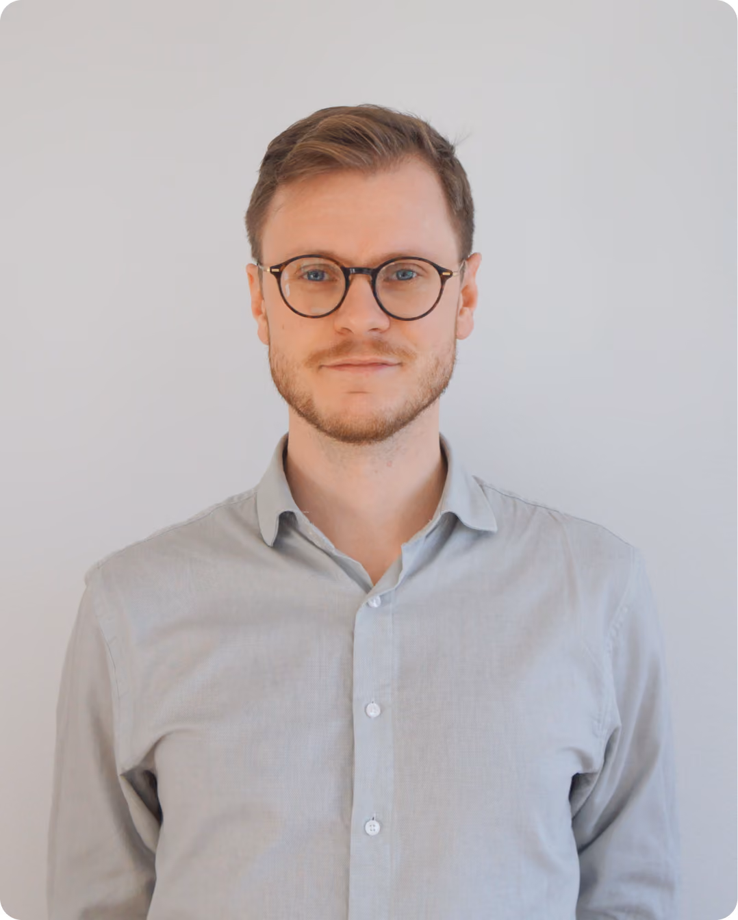 Man with light brown hair and beard wearing round glasses and a light gray button-up shirt.