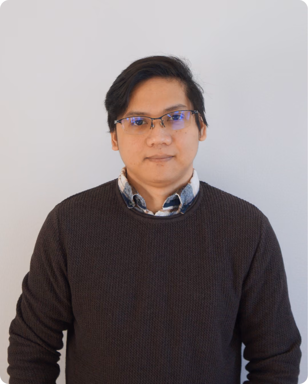 Man with glasses wearing a dark sweater over a checked shirt standing against a plain light background.