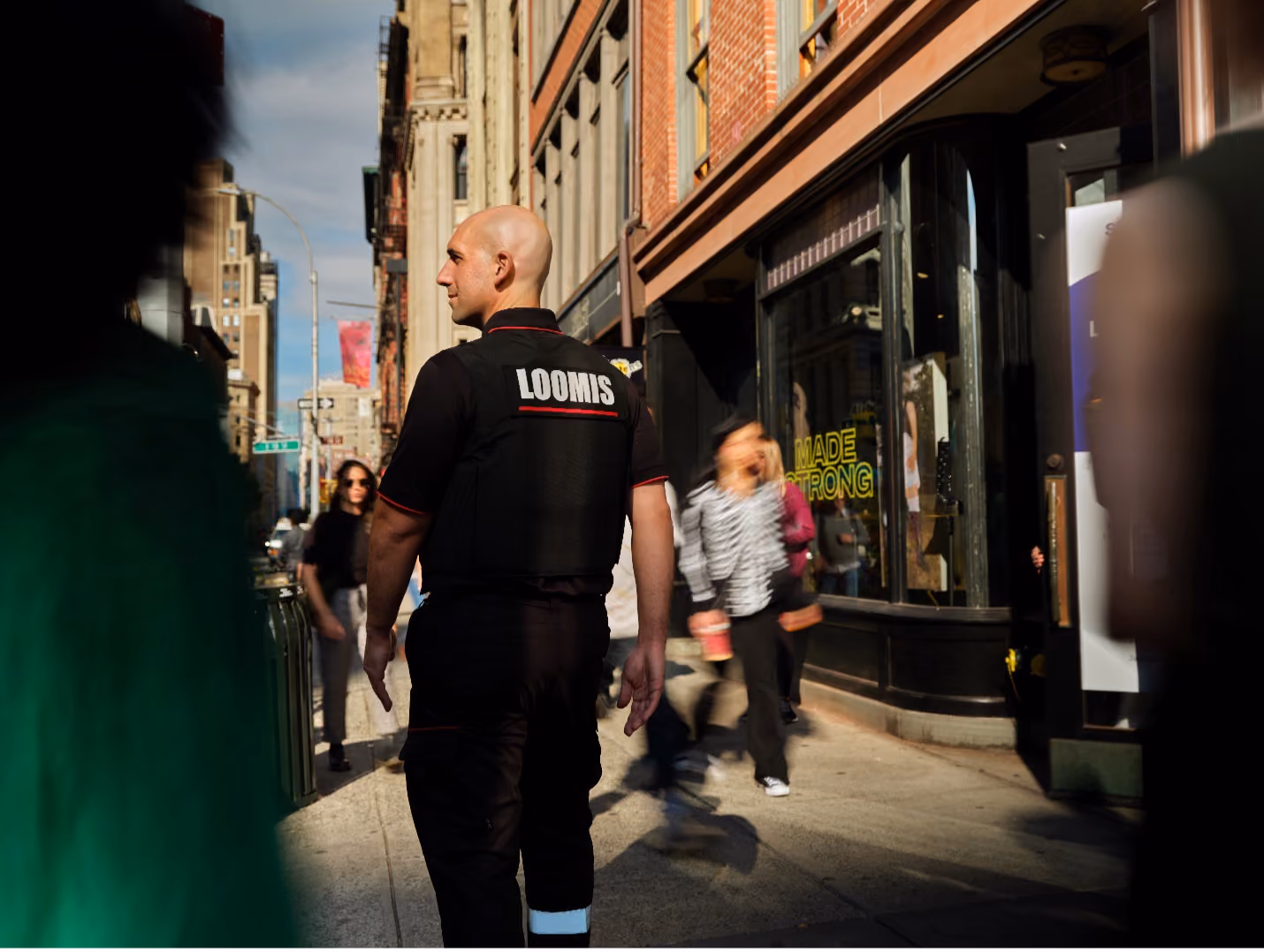Security guard wearing a vest labeled LOOMIS walking on a sunny city sidewalk with blurred pedestrians.