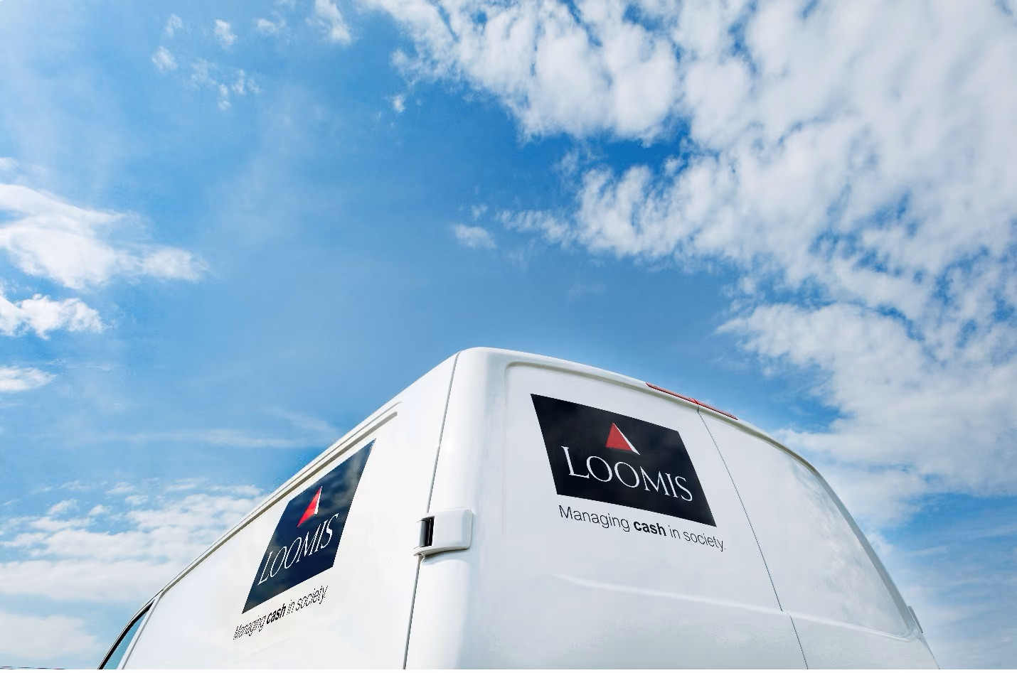 White van with Loomis logo and text 'Managing cash in society' against a blue sky with clouds.