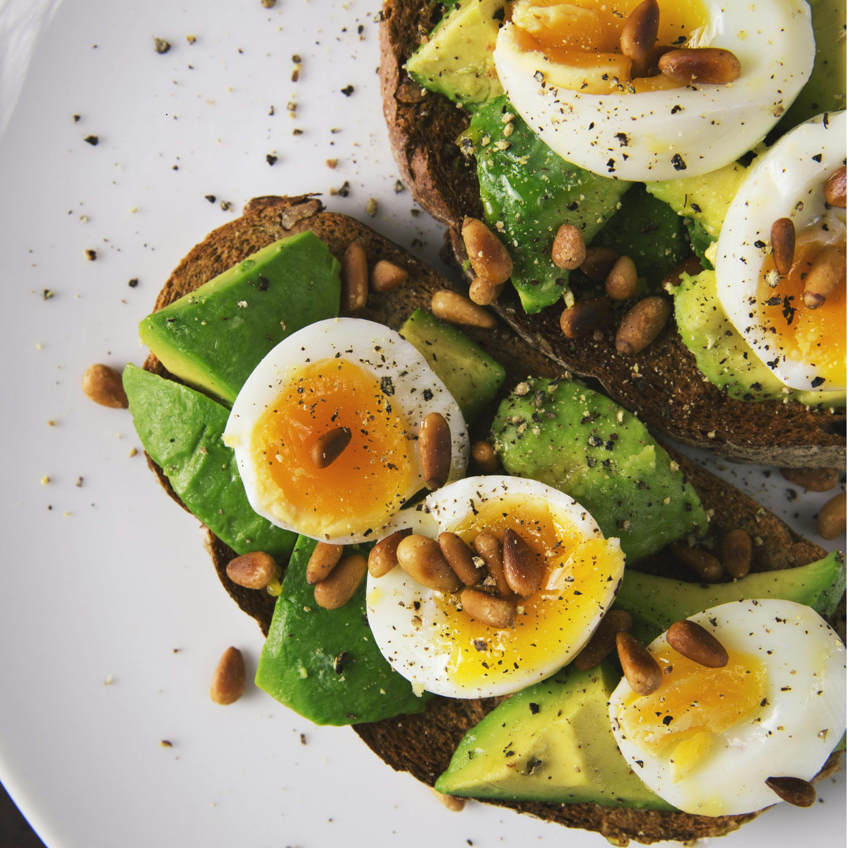 Toasted bread topped with sliced avocado, halved soft-boiled eggs, pine nuts, and black pepper on a white plate.