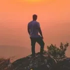 Person standing on a rocky hill overlooking a hazy landscape during an orange sunset.