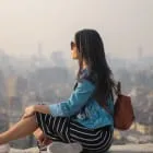 Woman in sunglasses and denim jacket sitting on a ledge overlooking a hazy cityscape.