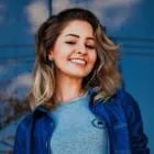 Smiling woman with shoulder-length wavy hair wearing a blue jacket and gray shirt outdoors.
