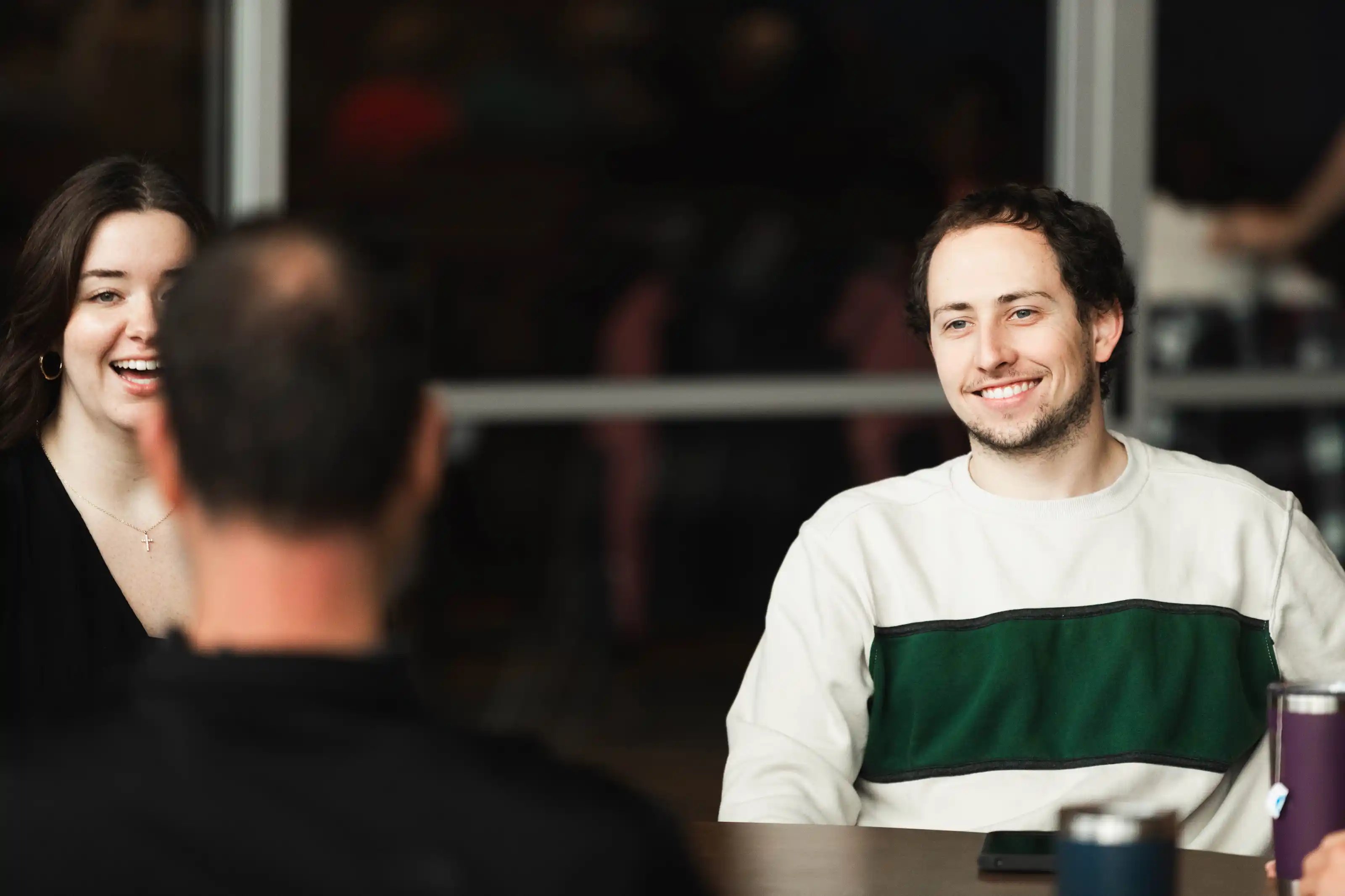 Two young adults smiling and talking across a table with a third person seen from behind.