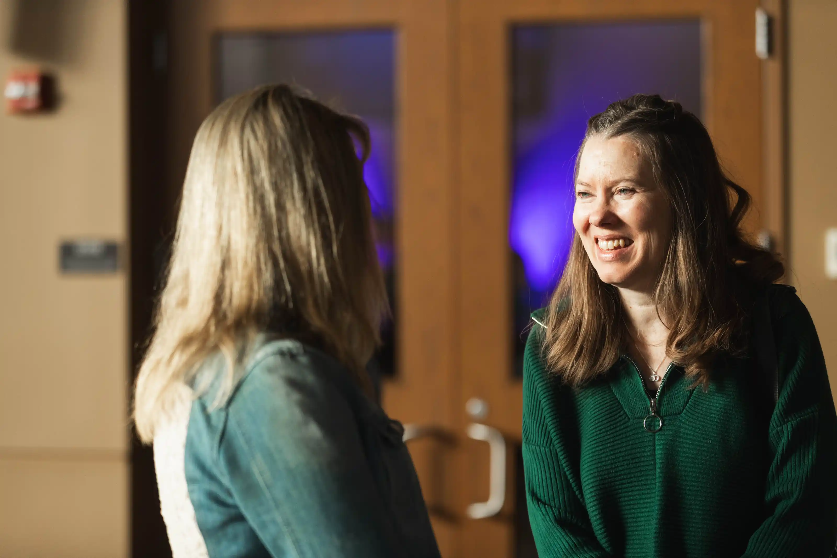 Two women engaged in conversation indoors, one smiling and wearing a green sweater.