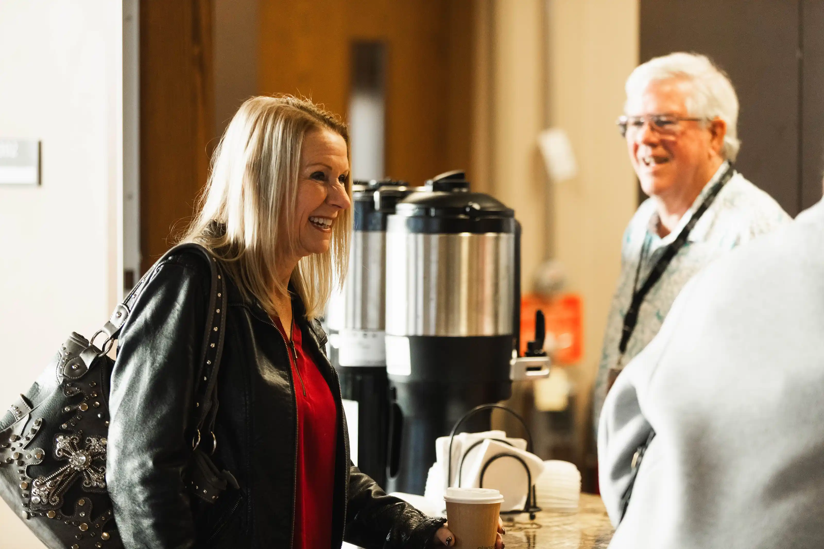 Woman with a coffee cup, smiling and conversing with a man near coffee dispensers.