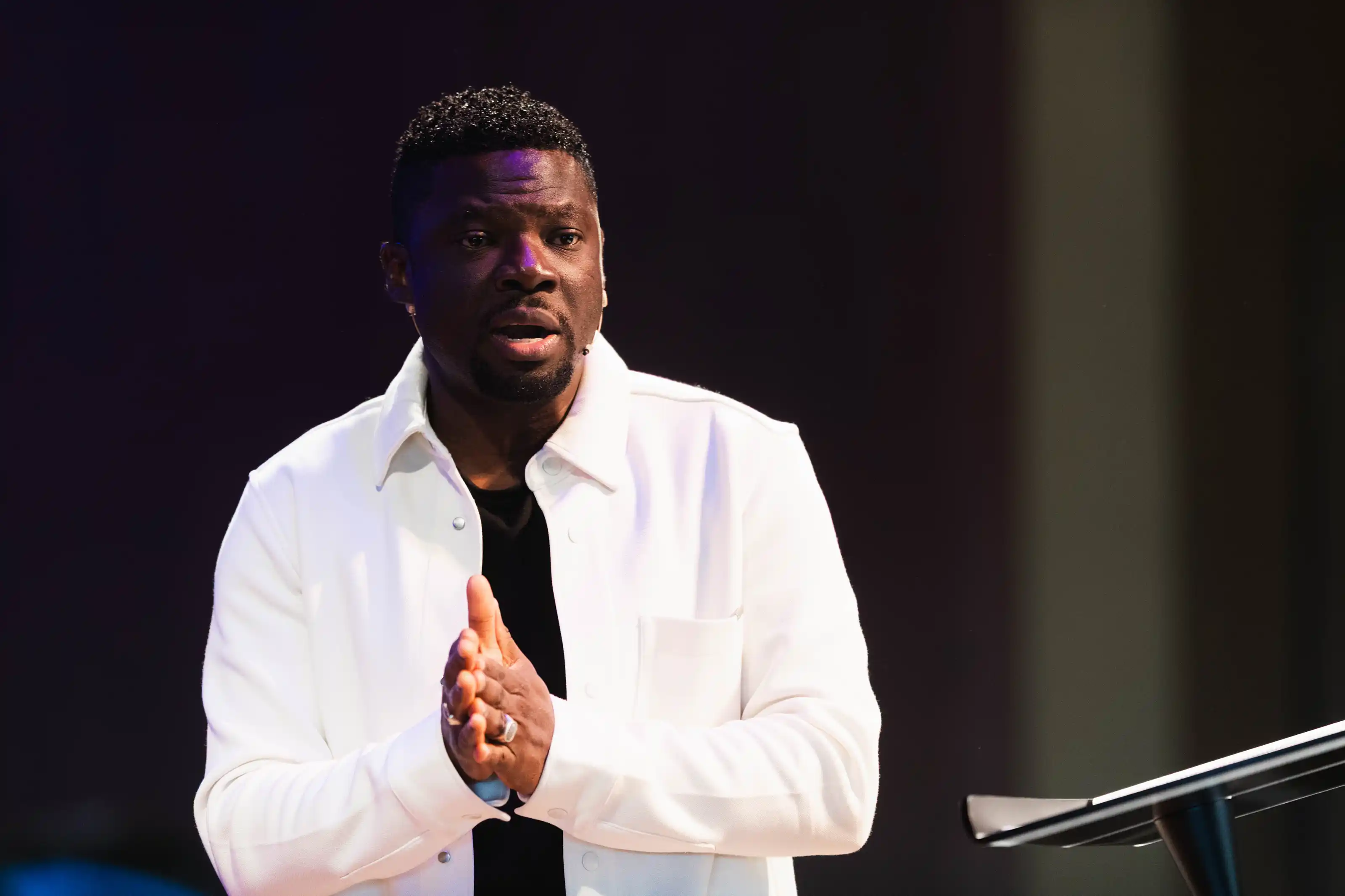 A man in a white jacket speaking or presenting with hands clasped in front of a dark background with a podium nearby.